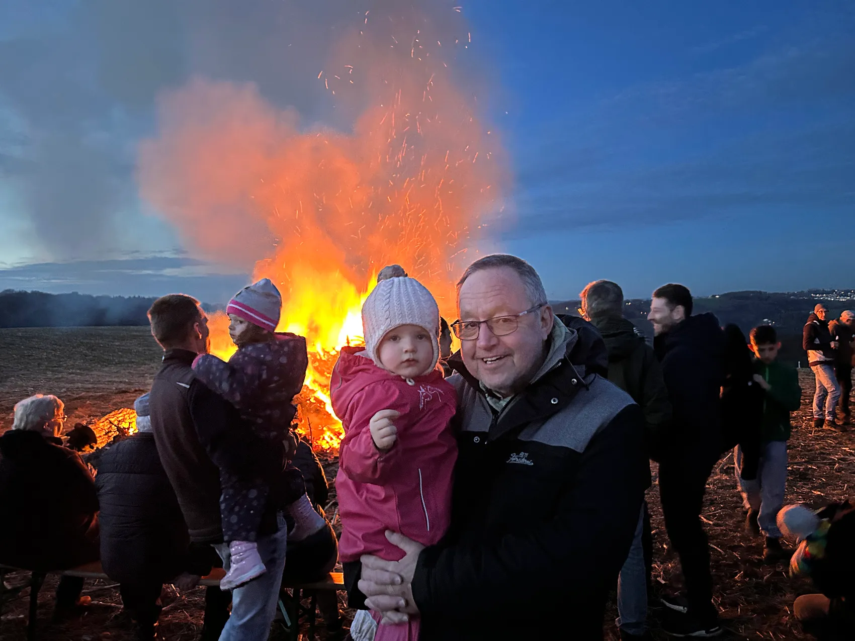Mann hält ein Kind auf dem Arm vor einem großen Osterfeuer, im Hintergrund weitere Menschen und ein dunkler Himmel.