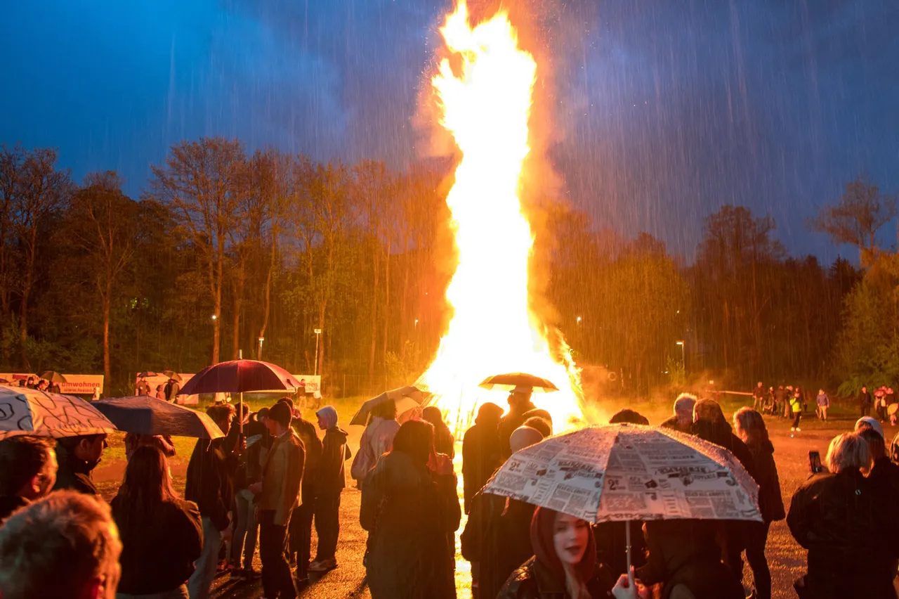 Großes Osterfeuer mit lodernden Flammen im Regen, umgeben von zahlreichen Zuschauern mit Regenschirmen.