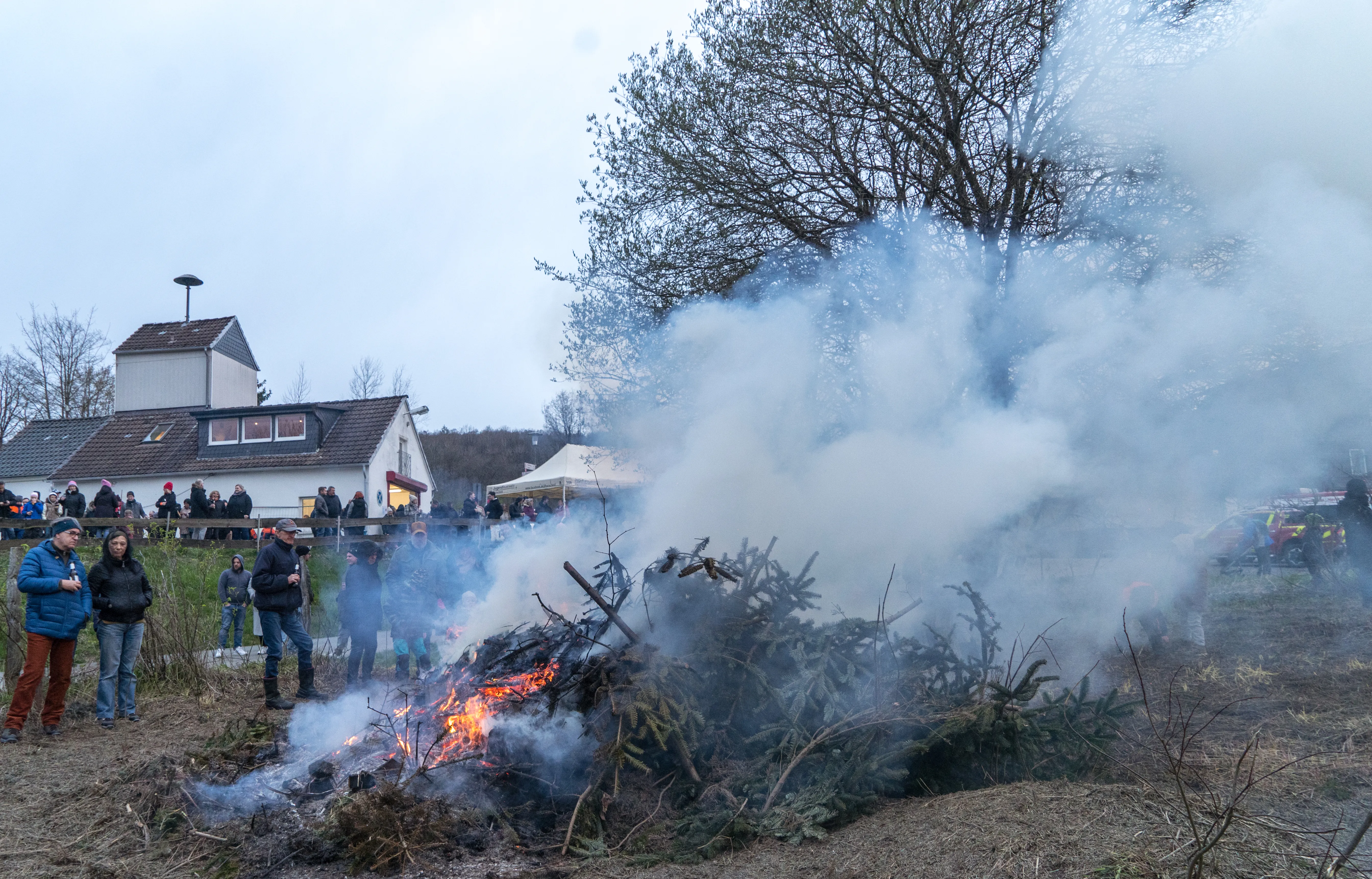 Großes Osterfeuer mit vielen Zuschauern, Rauch steigt auf. Im Hintergrund ein Wohnhaus und Bäume.