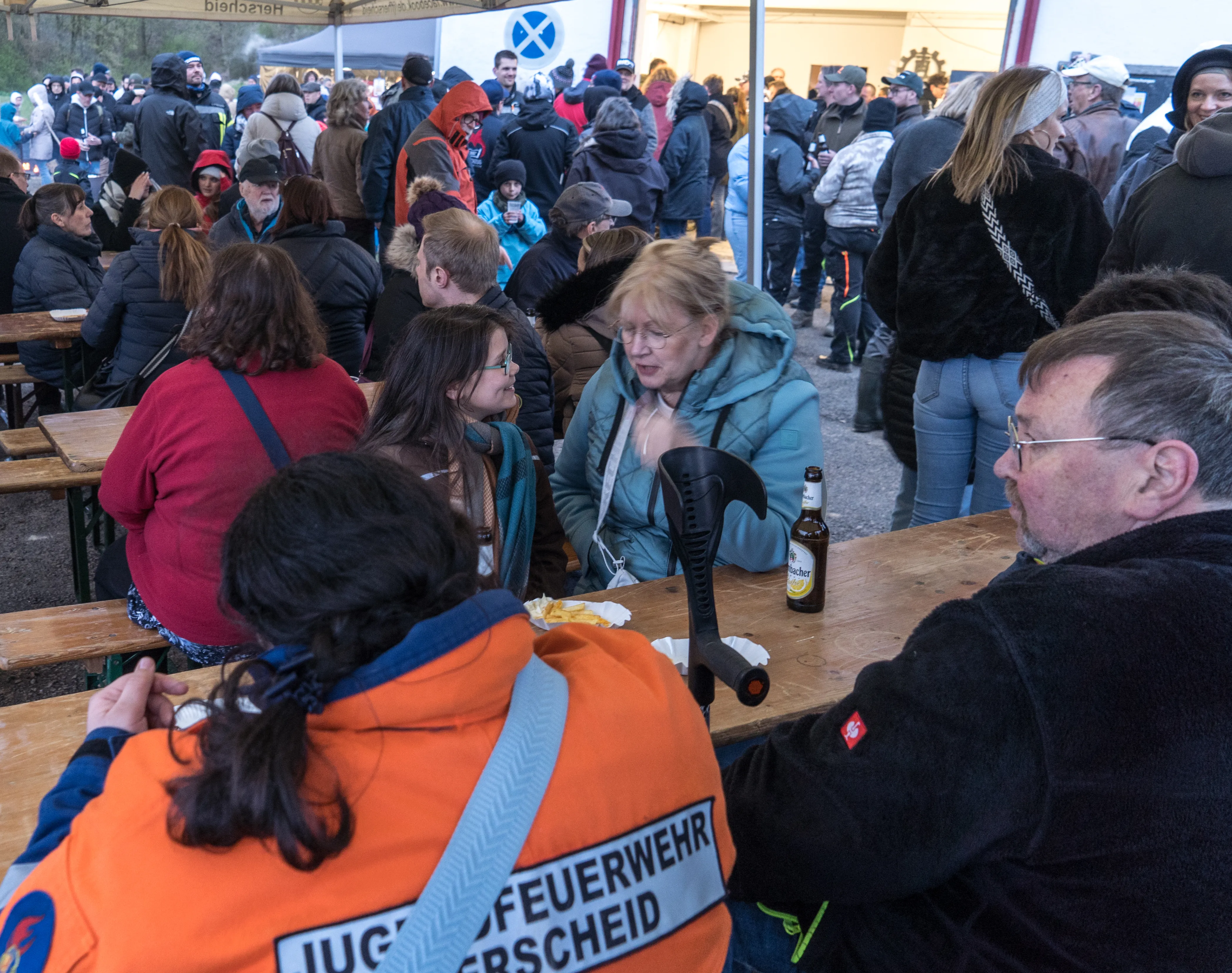 Menschen sitzen an Biertischbänken und unterhalten sich während eines Osterfeuers, im Vordergrund eine Person mit einer Jacke der Jugendfeuerwehr Herscheid.