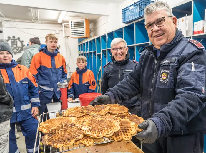 Ein Feuerwehrmann in Uniform hält ein Tablett mit vielen frisch gebackenen Waffeln vor eine Gruppe Jugendlicher.