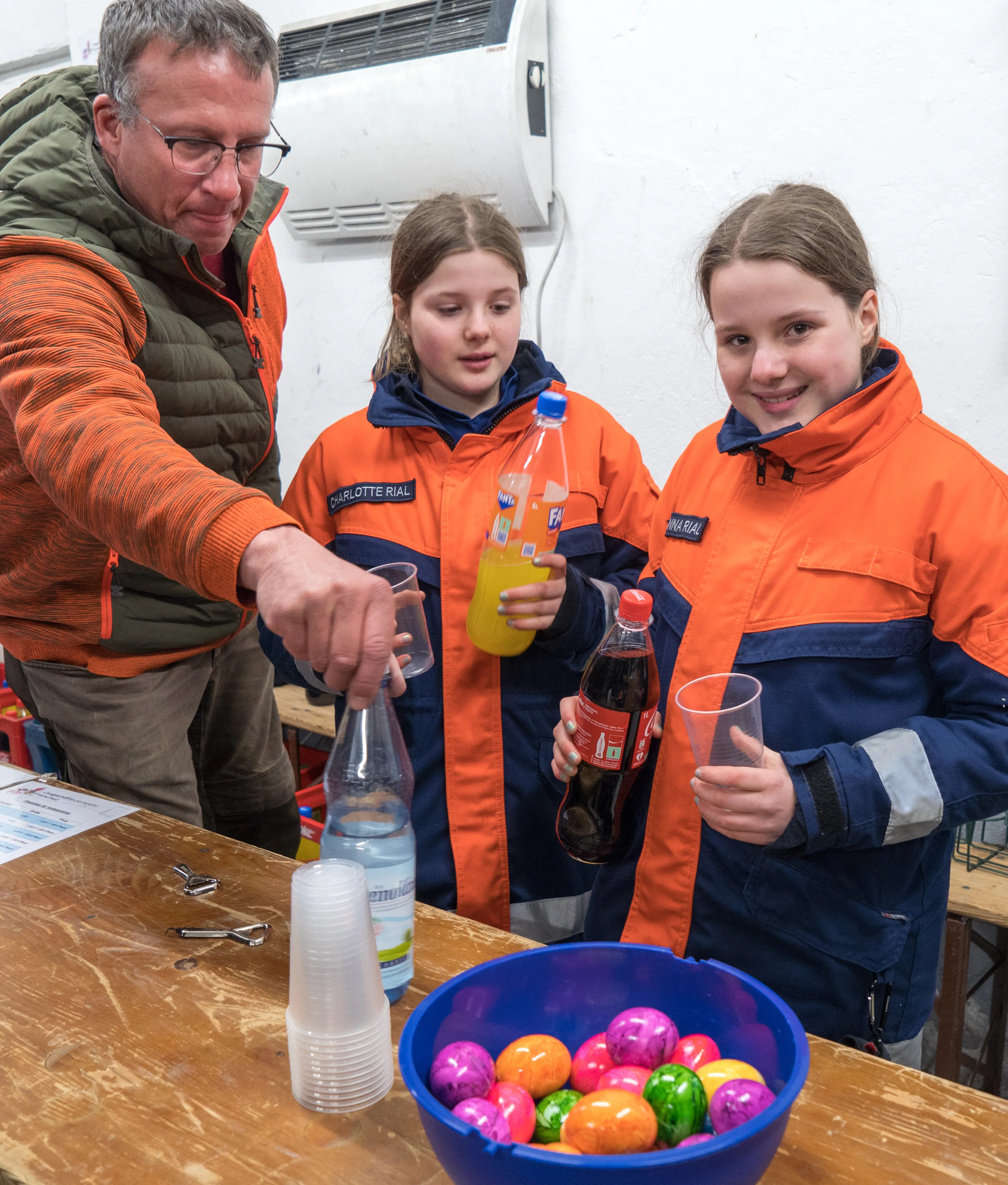 Ein Mann der Jugendfeuerwehr füllt Getränke in Gläser für zwei Mädchen in orangefarbenen Jacken. Vor ihnen steht eine Schüssel mit bunten Ostereiern.