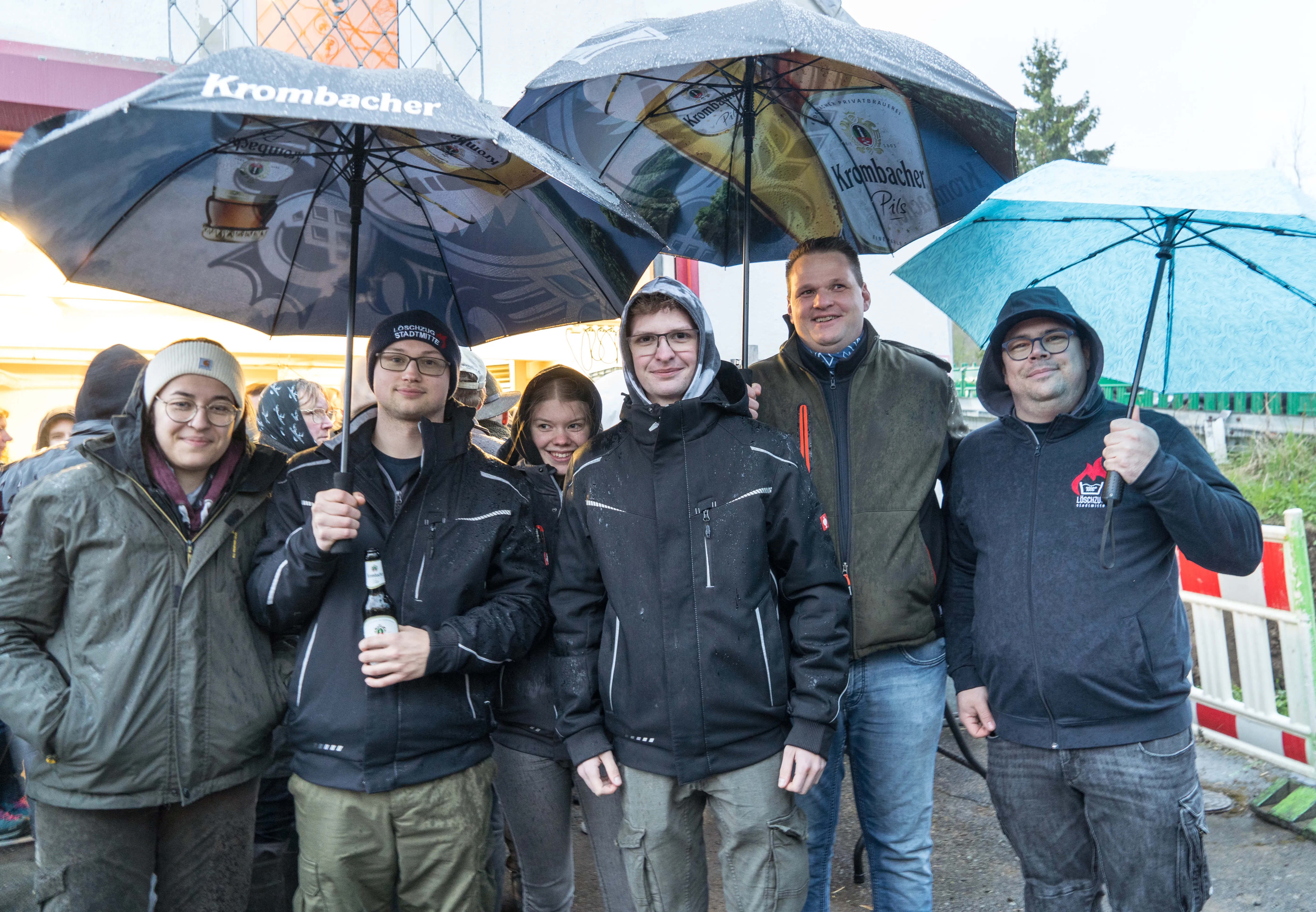 Sechs junge Erwachsene der Jugendfeuerwehr stehen lächelnd unter Schirmen mit Werbedruck für Krombacher Pils zusammen. Im Hintergrund sind weitere Menschen und ein Absperrgitter zu sehen.
