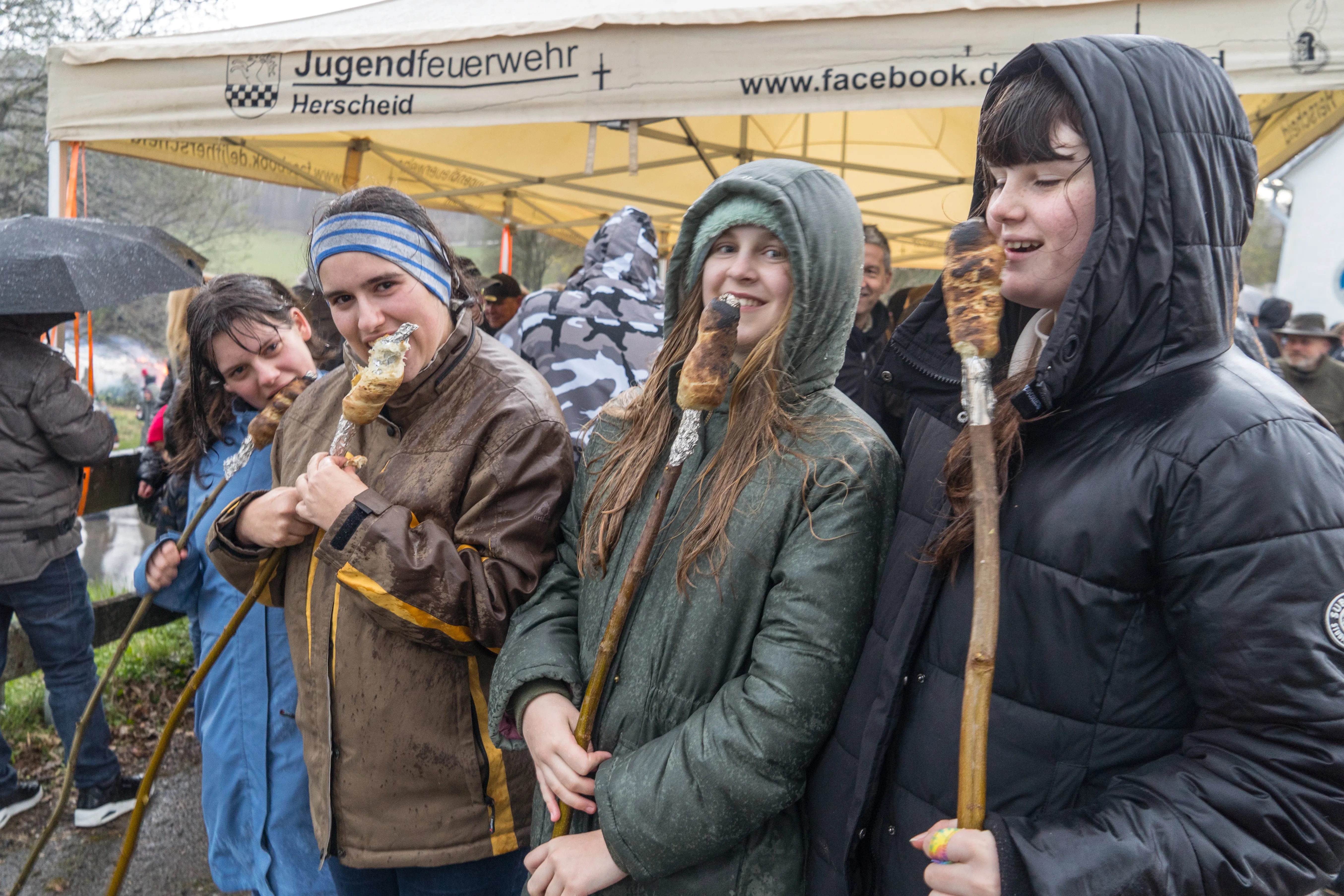 Drei Jugendliche halten jeweils einen mit Teig umwickelten Stock über offenem Feuer fest. Im Hintergrund ein Zelt der Jugendfeuerwehr Herscheid und weitere Menschen.