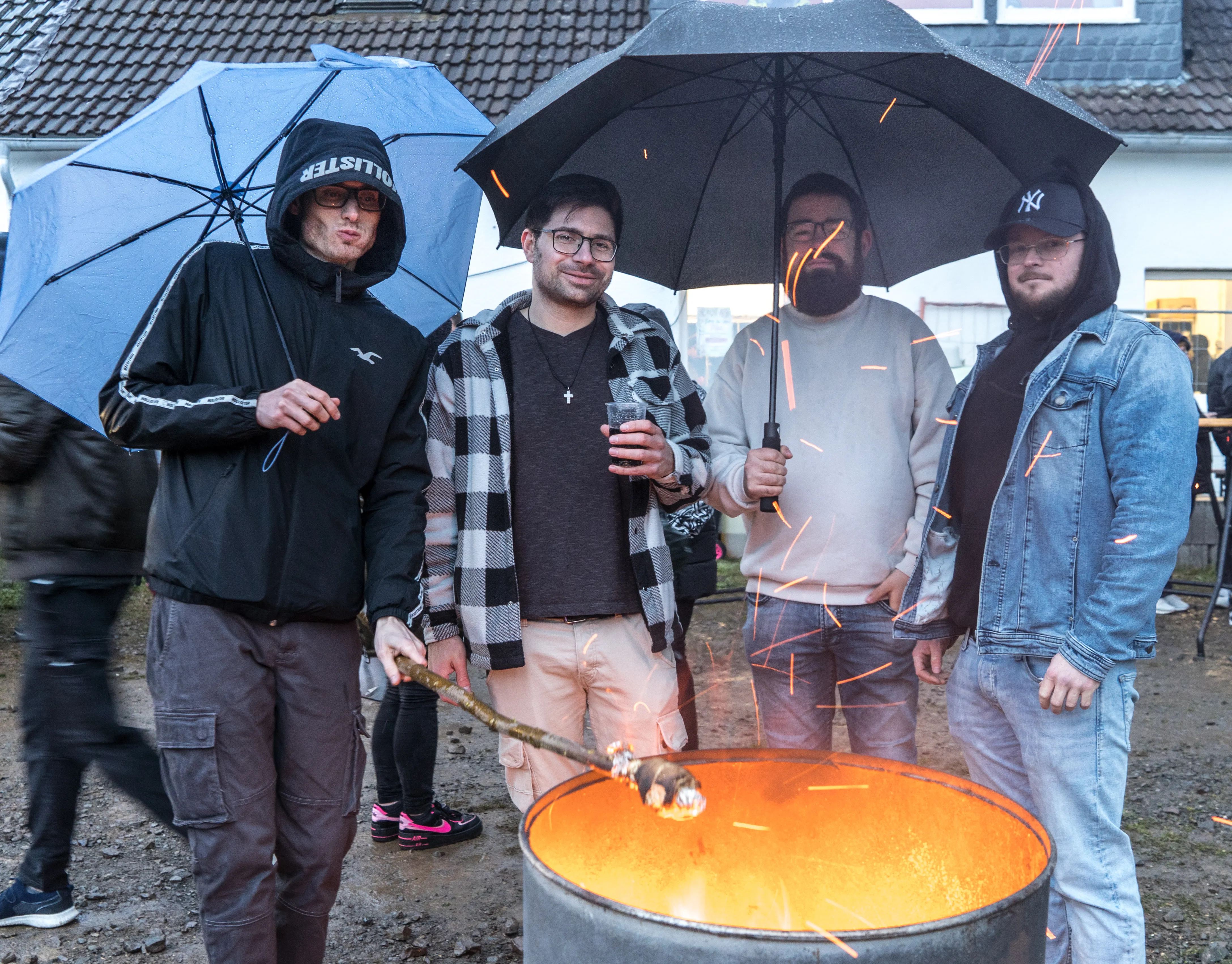 Vier junge Männer stehen um ein großes Feuer in einer Metallschale, geschützt durch Regenschirme. Im Hintergrund sind weitere Personen und Häuser zu sehen.