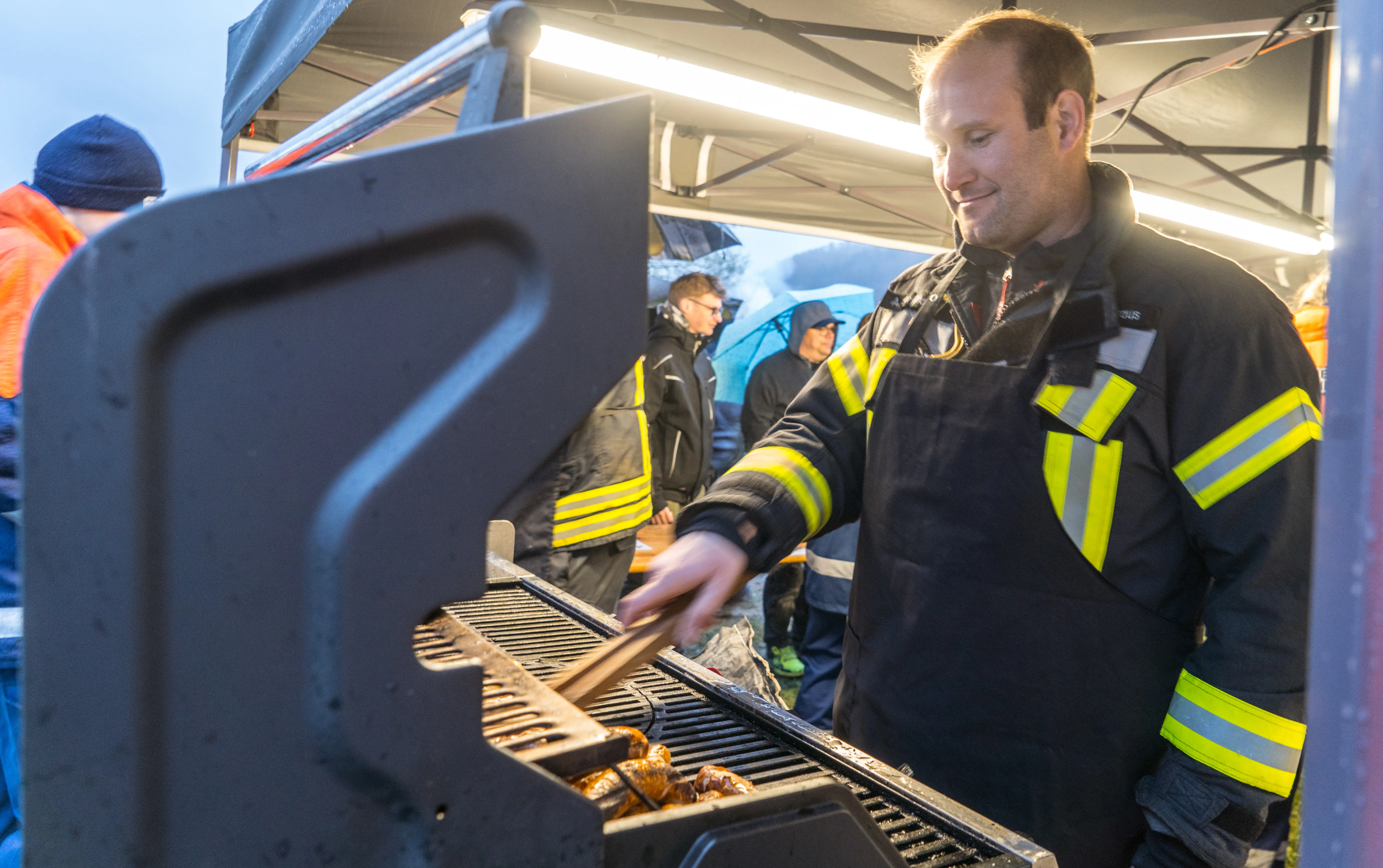 Ein Feuerwehrmann grillt Fleisch auf einem Holzkohlegrill unter einem Zelt, während im Hintergrund weitere Feuerwehrleute stehen.