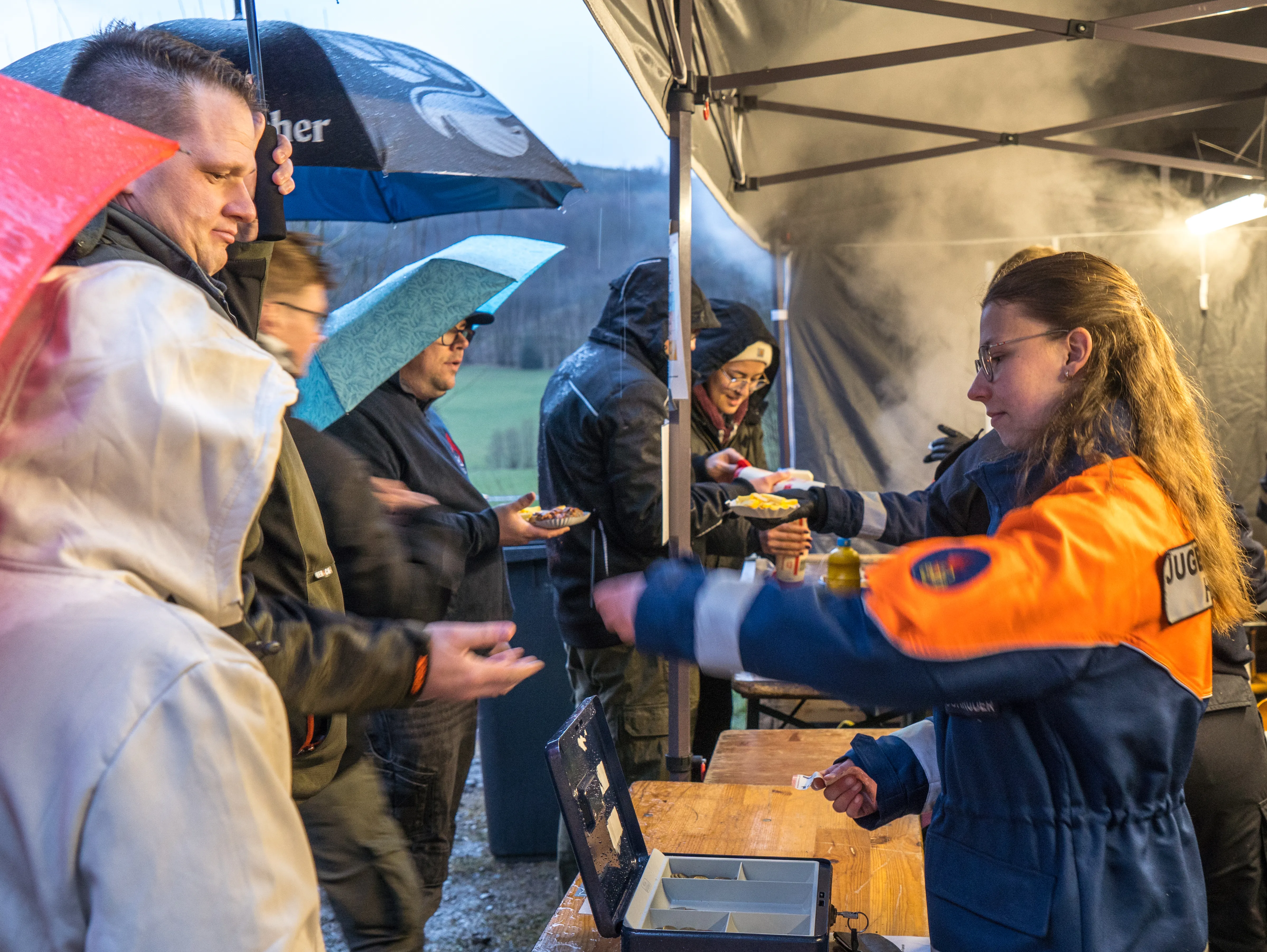Eine Frau in Uniform der Jugendfeuerwehr reicht unter einem Zelt Essen an eine Schlange wartender Personen weiter, während es regnet.
