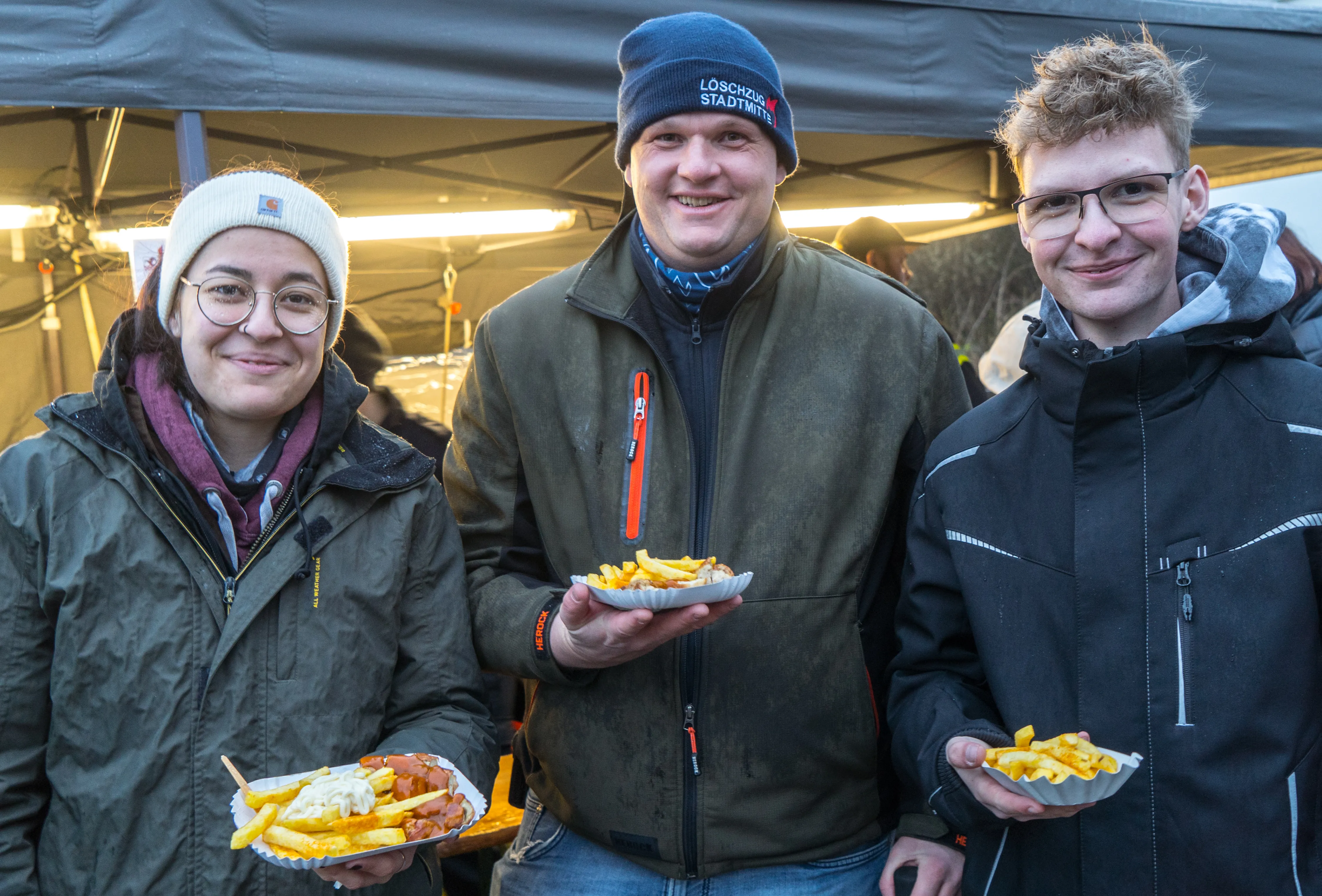 Drei junge Erwachsene halten jeweils eine Portion Pommes oder Pommes mit Currywurst vor sich. Im Hintergrund ein dunkler Zeltaufbau.