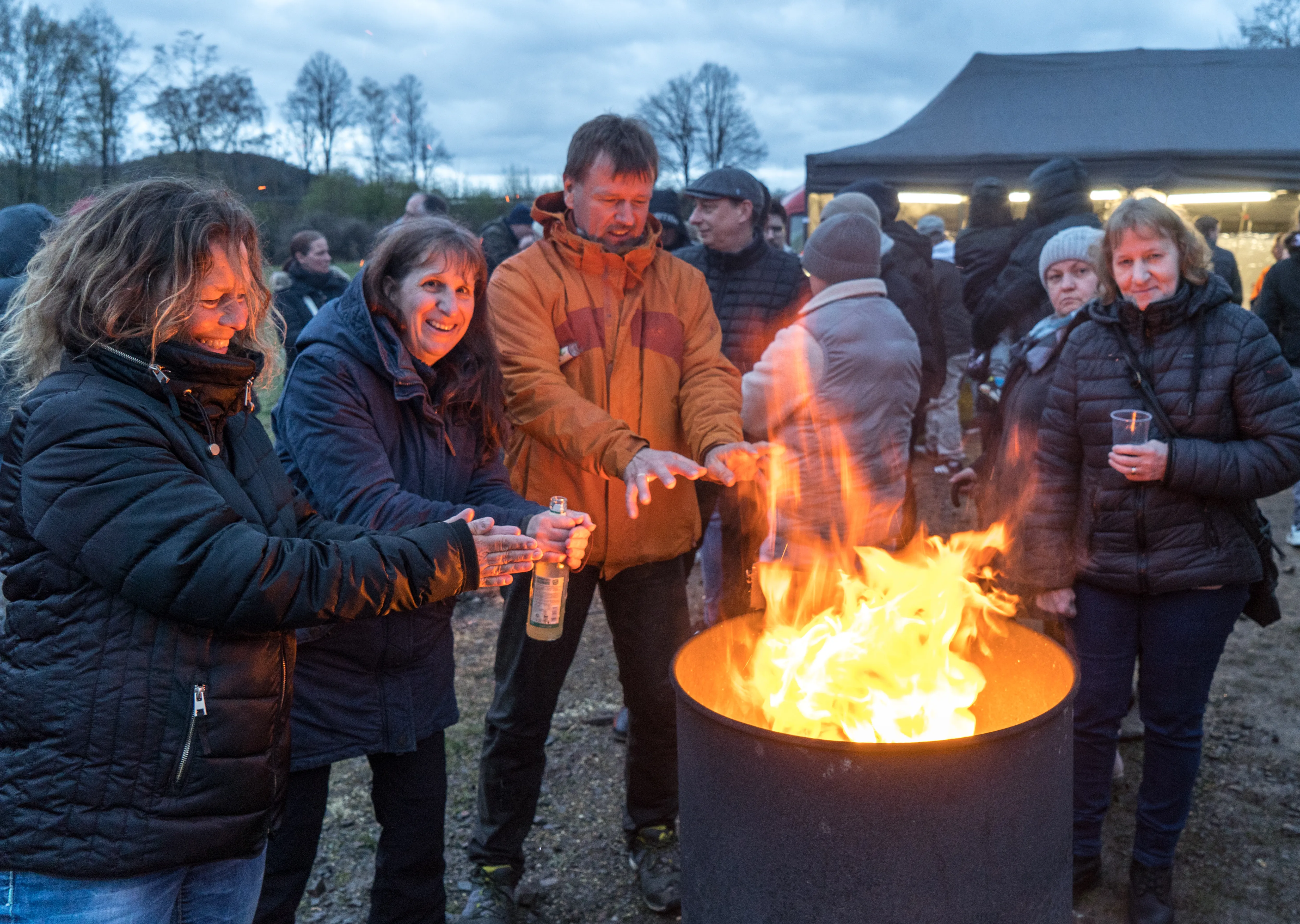 Menschen stehen um ein großes Feuer in einer Stahltonne herum, einige halten Getränke in der Hand. Im Hintergrund sind weitere Personen und ein Zelt zu sehen.