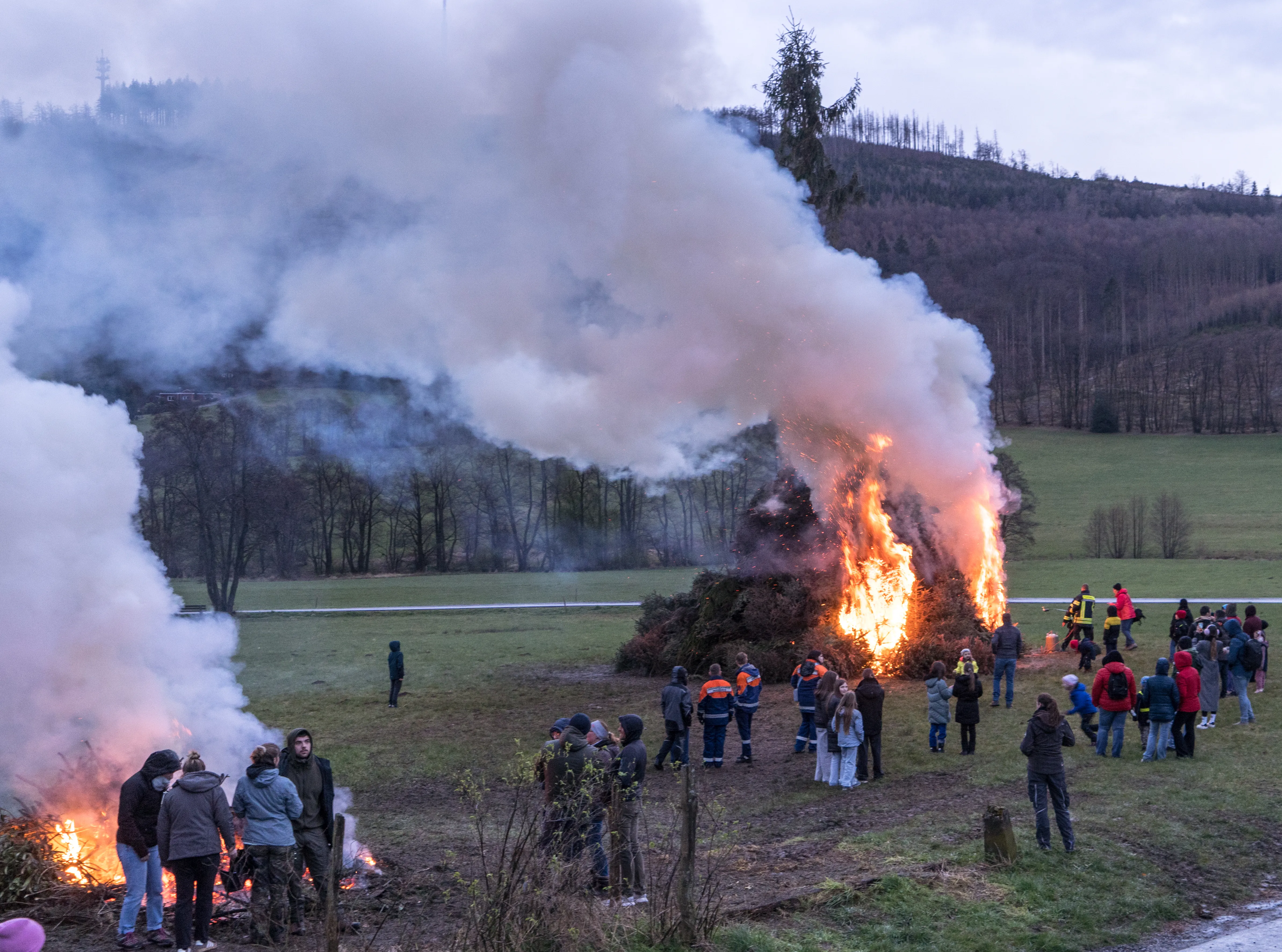 Großes Osterfeuer mit viel Rauch auf einer Wiese, umgeben von zahlreichen Zuschauern, darunter Mitglieder der Jugendfeuerwehr in orangefarbenen Westen.