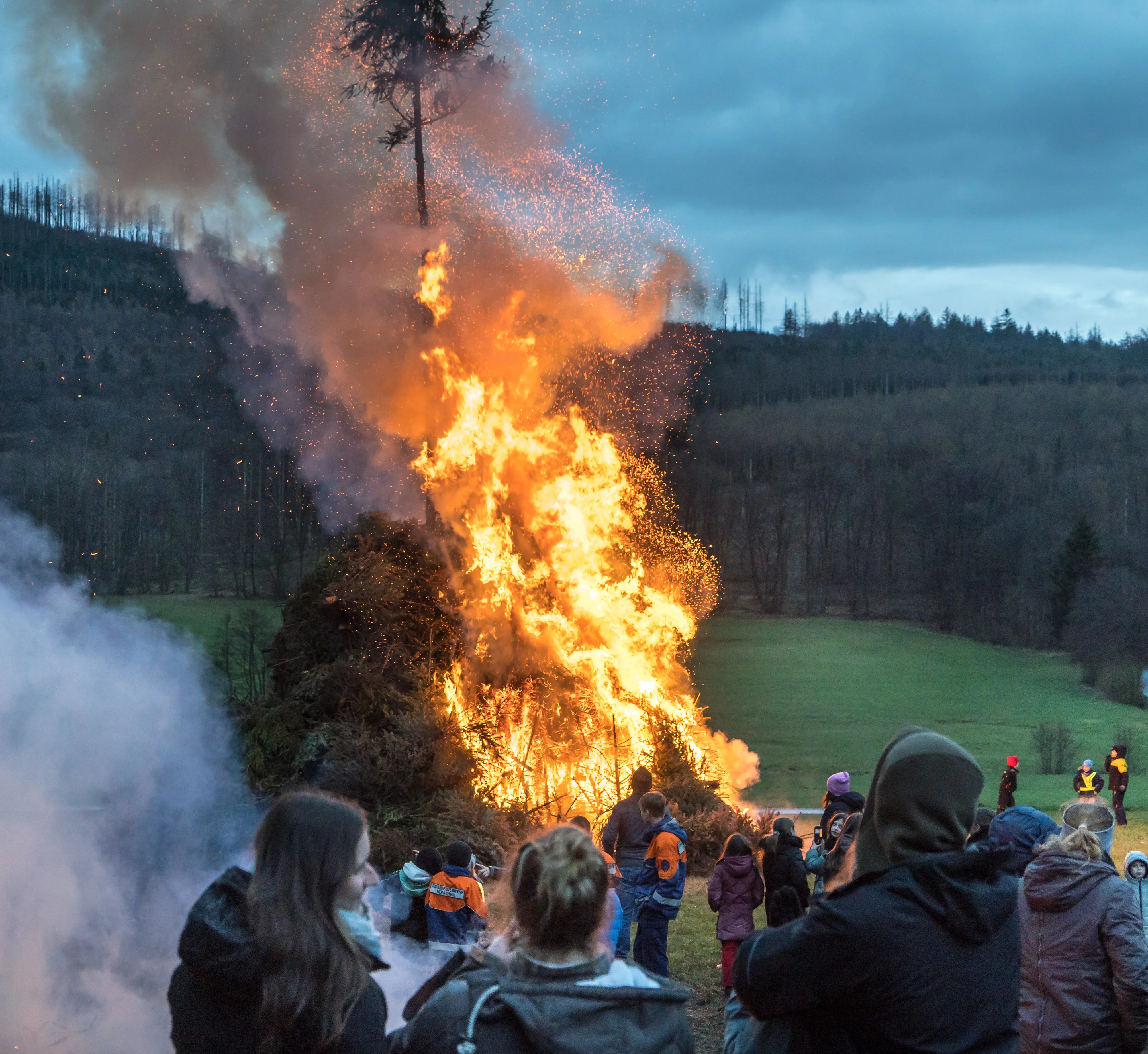 Großes Osterfeuer mit vielen Zuschauern, Rauch und Funken steigen auf. Im Hintergrund eine bewaldete Hügellandschaft.