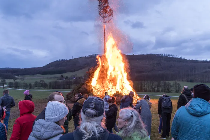 Großes Osterfeuer mit einer hohen Flammenzunge und Funkenflug, umgeben von einer Menschenmenge auf einer Wiese vor bewaldeten Hügeln.