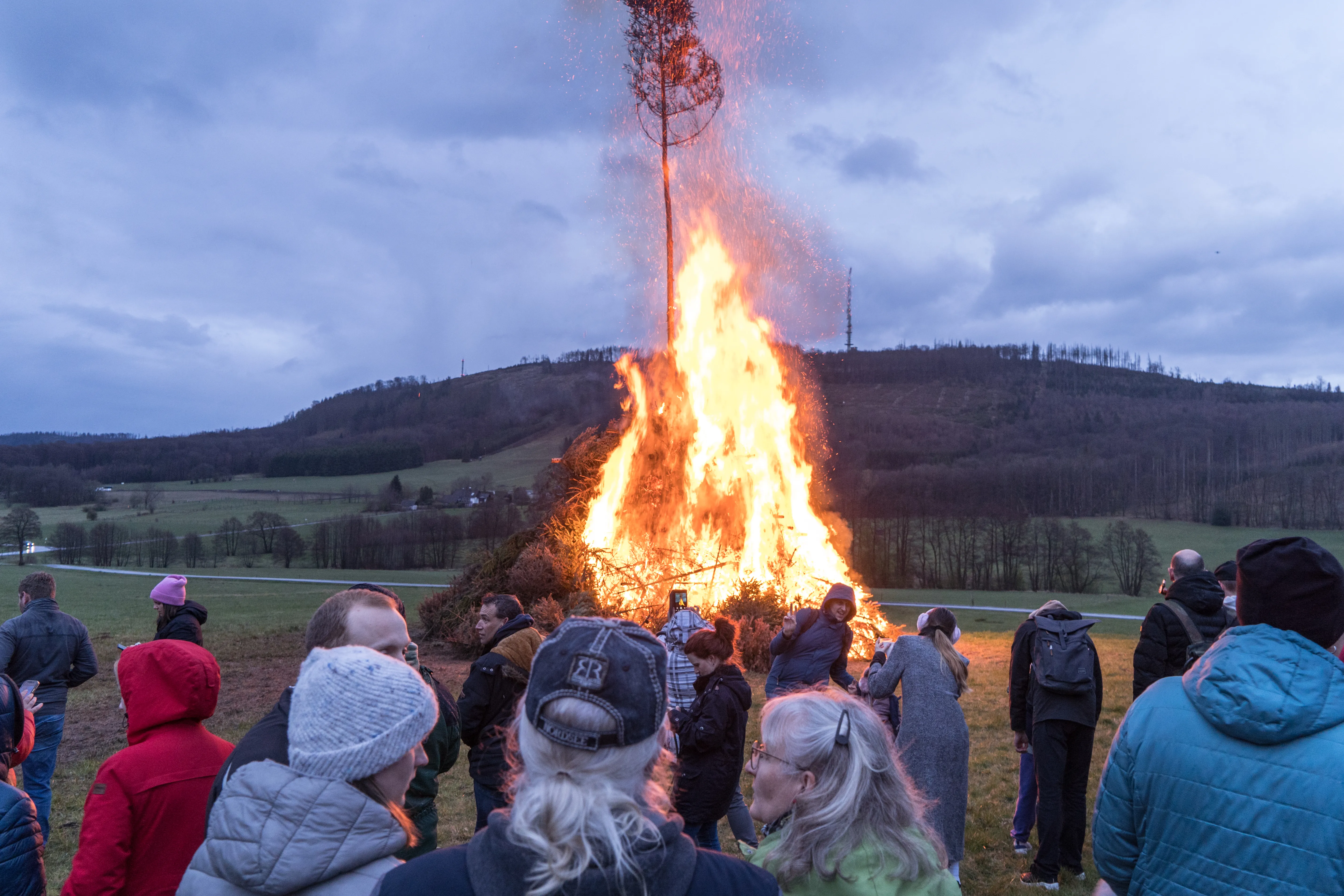 Großes Osterfeuer mit einer hohen Flammenzunge und Funkenflug, umgeben von einer Menschenmenge auf einer Wiese vor bewaldeten Hügeln.