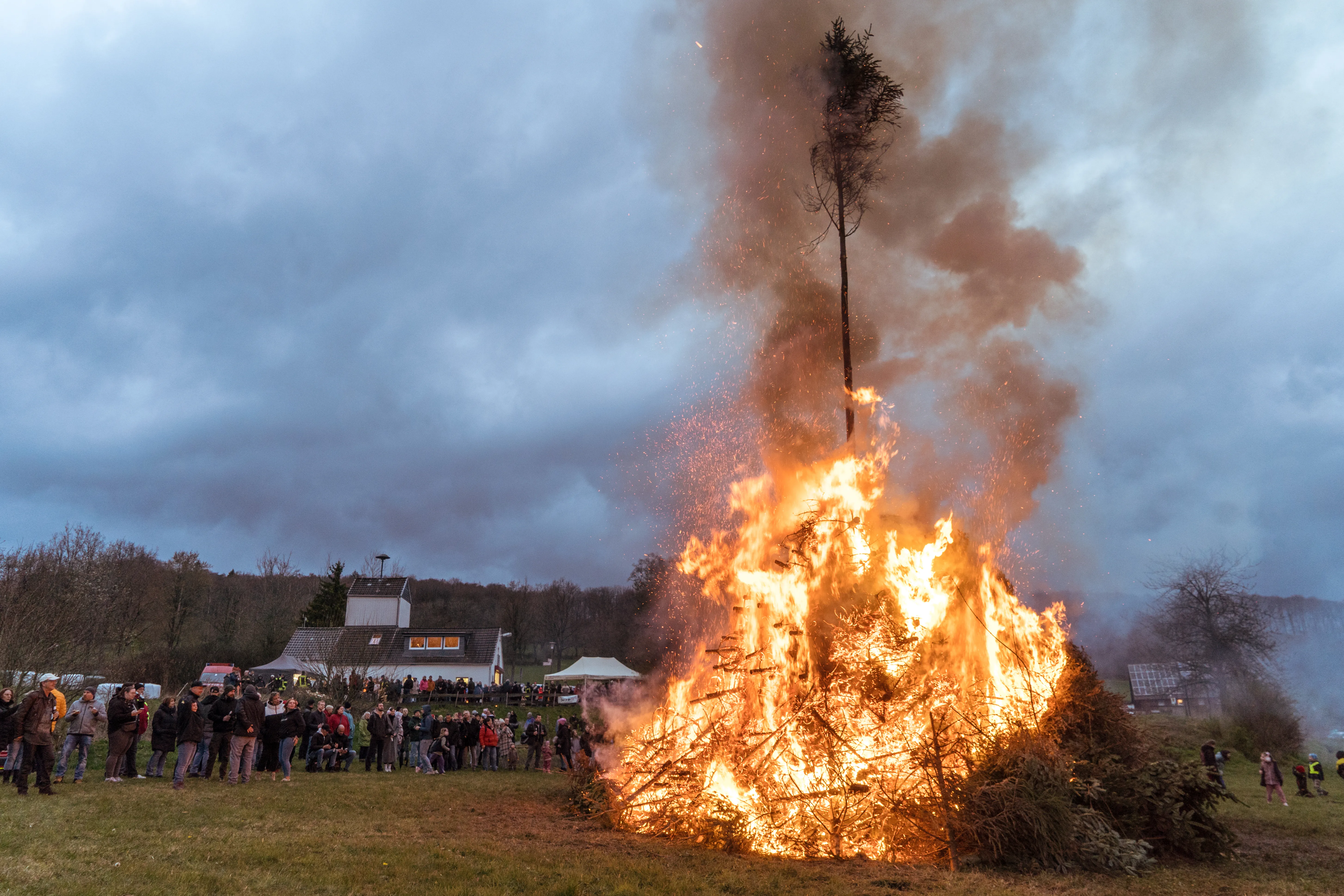Großes Osterfeuer mit hohem Holzstapel und lodernden Flammen, umgeben von einer Menschenmenge und im Hintergrund einem Gebäude.