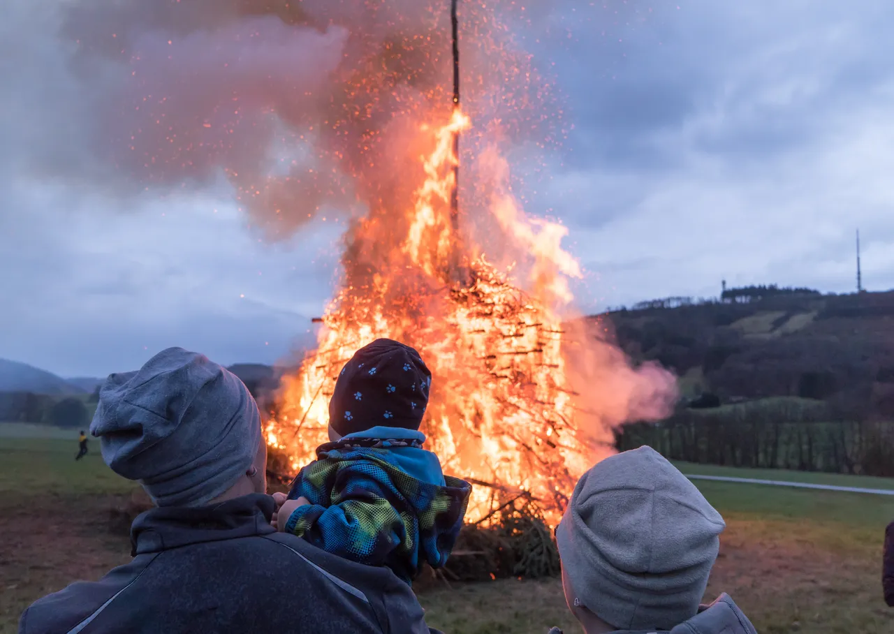 Drei Personen, darunter ein Kind auf den Schultern, blicken auf ein großes Osterfeuer mit hohen Flammen und Funkenflug. Im Hintergrund eine Hügellandschaft.
