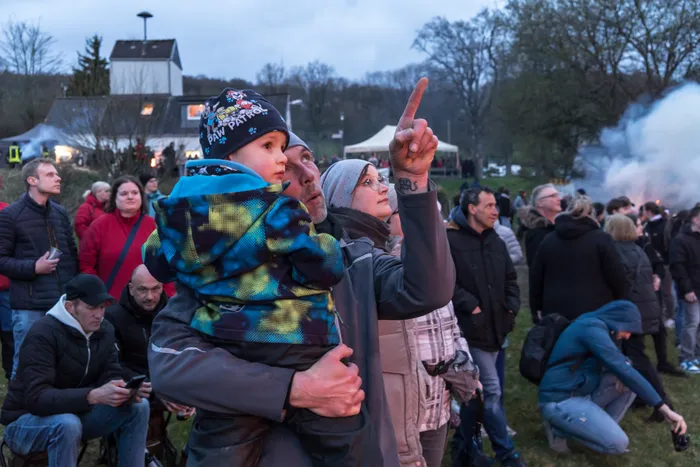Zuschauer beobachten ein großes Osterfeuer, während eine Frau mit einem Kind auf den Schultern in den Himmel zeigt. Im Hintergrund ist ein Gebäude und Rauch zu sehen.