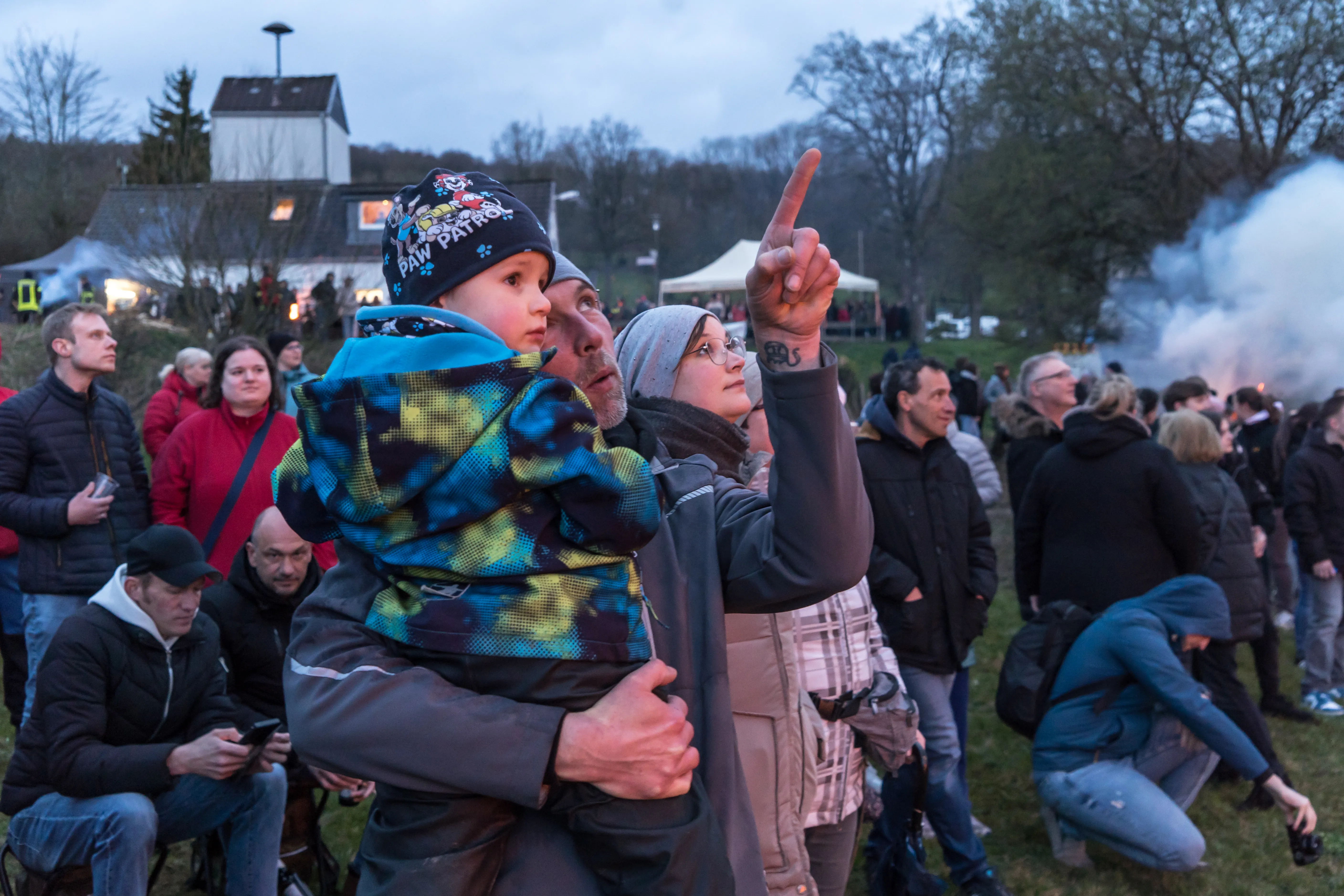 Zuschauer beobachten ein großes Osterfeuer, während eine Frau mit einem Kind auf den Schultern in den Himmel zeigt. Im Hintergrund ist ein Gebäude und Rauch zu sehen.