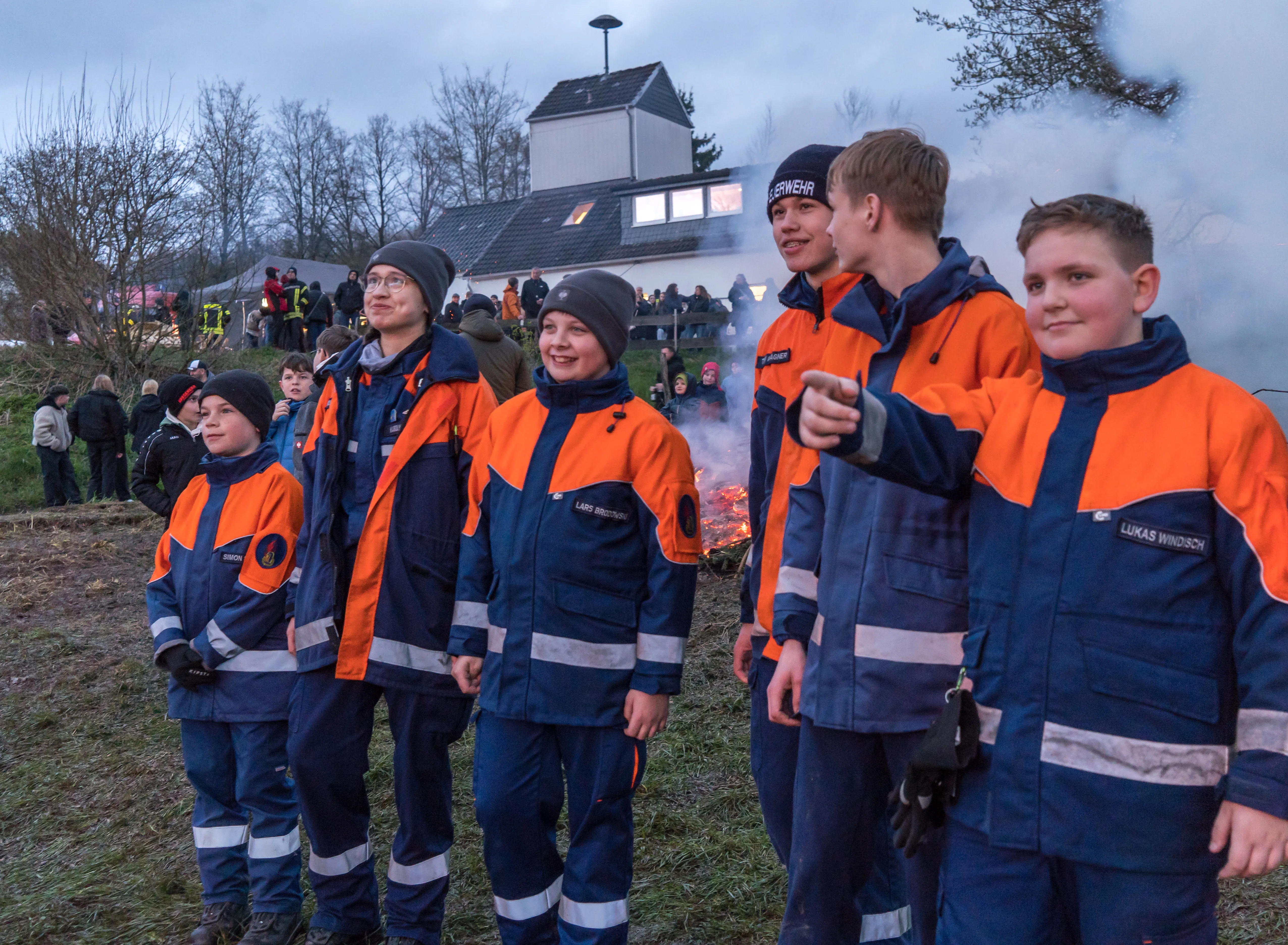 Jugendfeuerwehrleute in orange-blauen Jacken stehen vor einem großen Osterfeuer, im Hintergrund ein Gebäude und weitere Menschen.