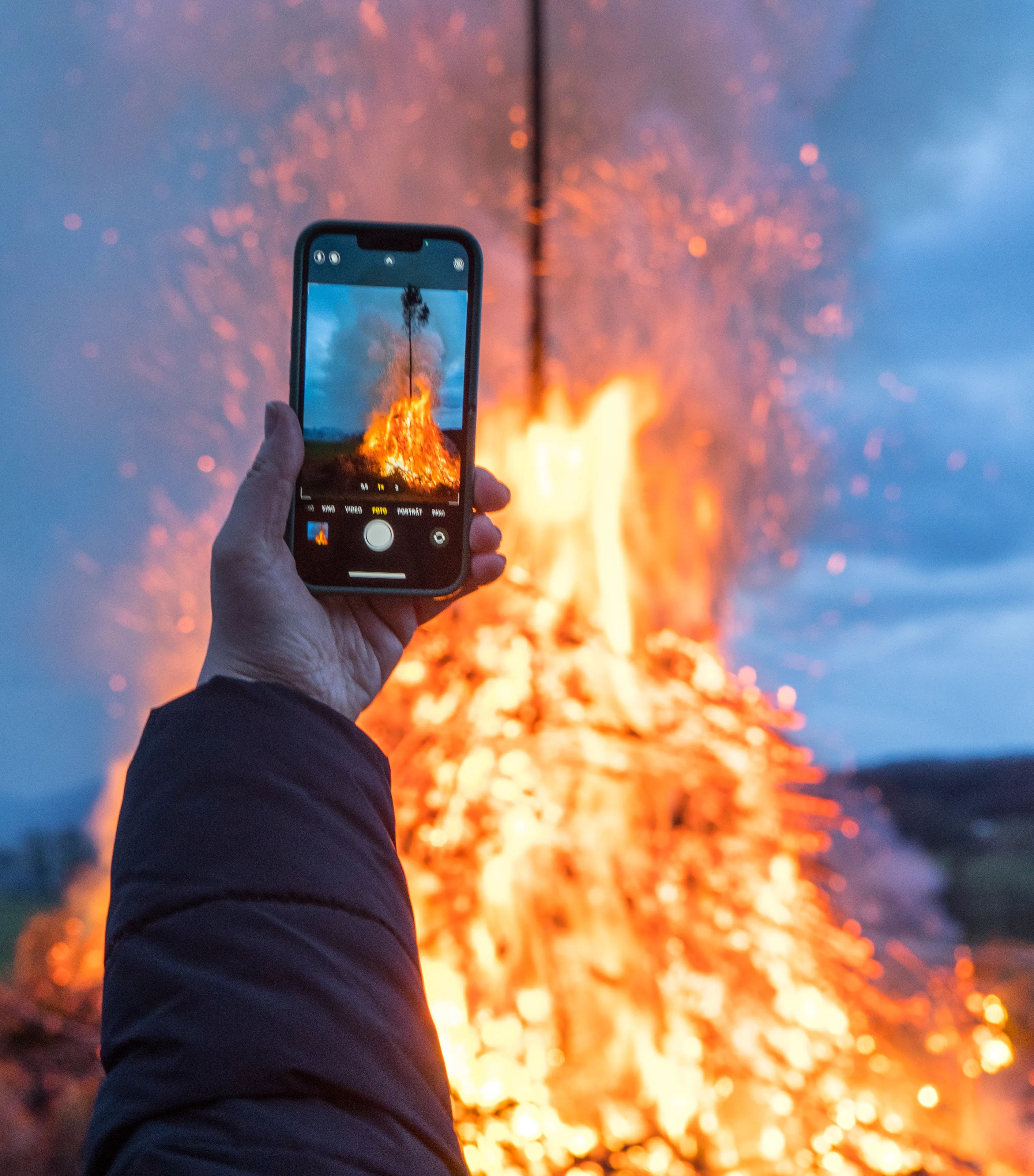 Eine Person filmt mit einem Smartphone ein großes Osterfeuer, das in den Abendhimmel aufsteigt. Im Hintergrund sind Funken und Rauch zu sehen.