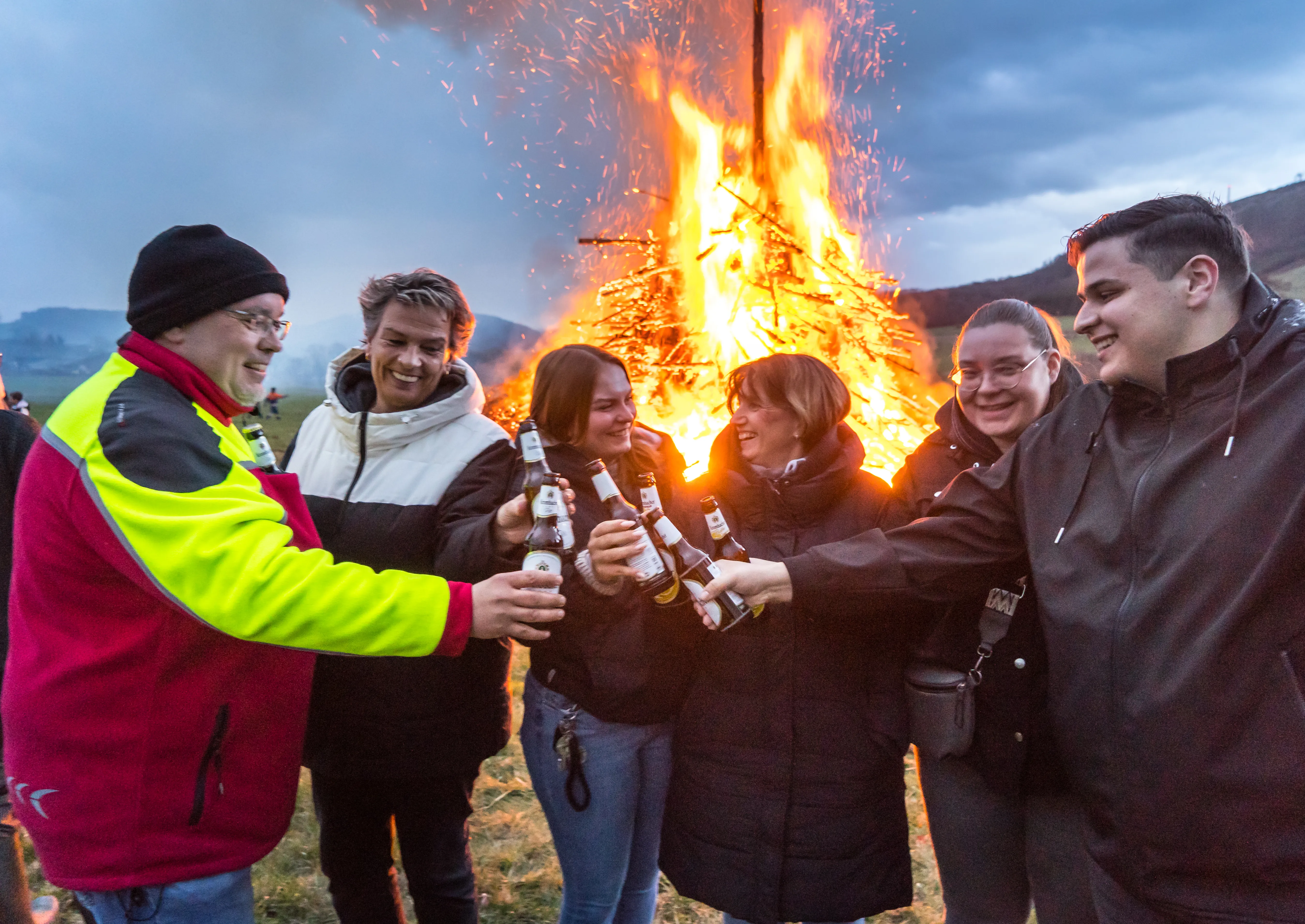 Eine Gruppe junger Erwachsener erhebt mit Bierflaschen vor einem großen Osterfeuer. Im Hintergrund eine bewaldete Hügellandschaft.
