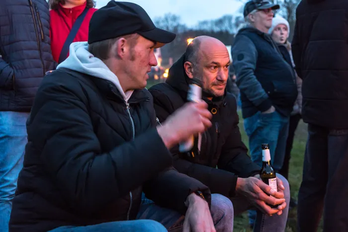 Zwei Männer sitzen nebeneinander und unterhalten sich während eines Osterfeuers, im Hintergrund weitere Personen und das brennende Feuer.