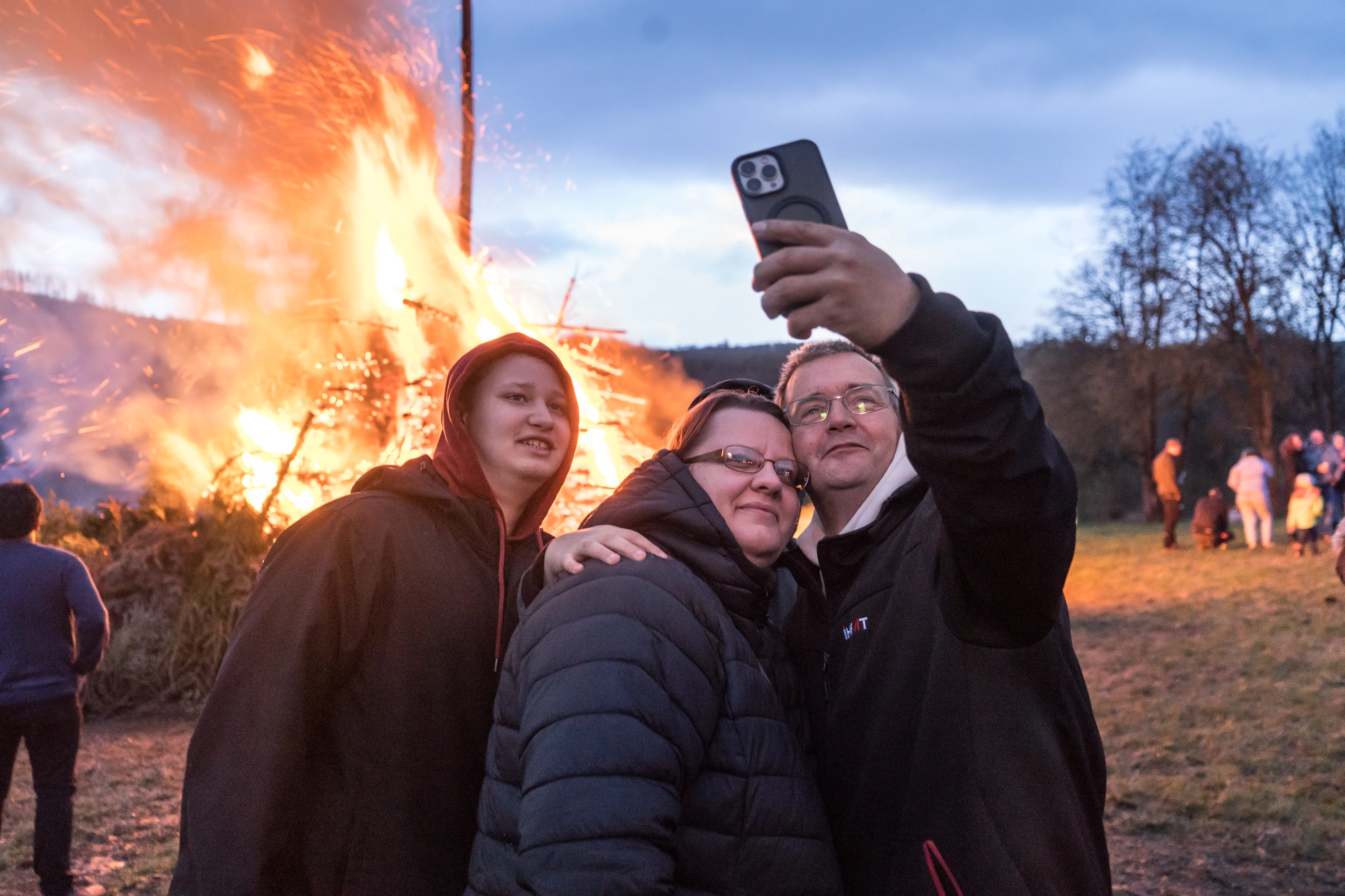 Drei Personen machen ein Selfie vor einem großen Osterfeuer mit Funkenflug, im Hintergrund verschwommene Menschen und Bäume.