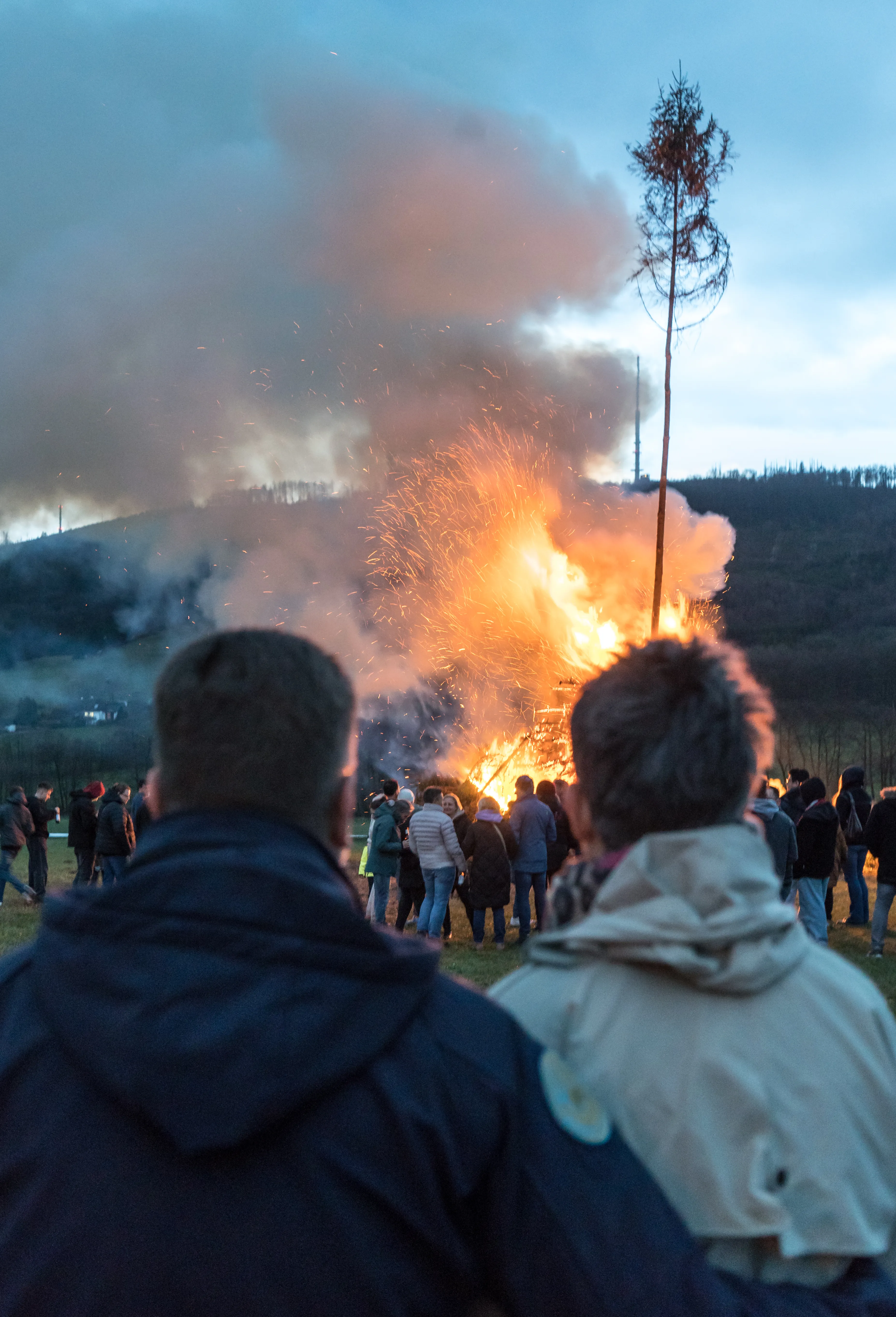 Großes Osterfeuer mit vielen Menschen im Hintergrund, Rauch steigt in den Himmel. Im Hintergrund eine Baumspitze und ein Sendemast.