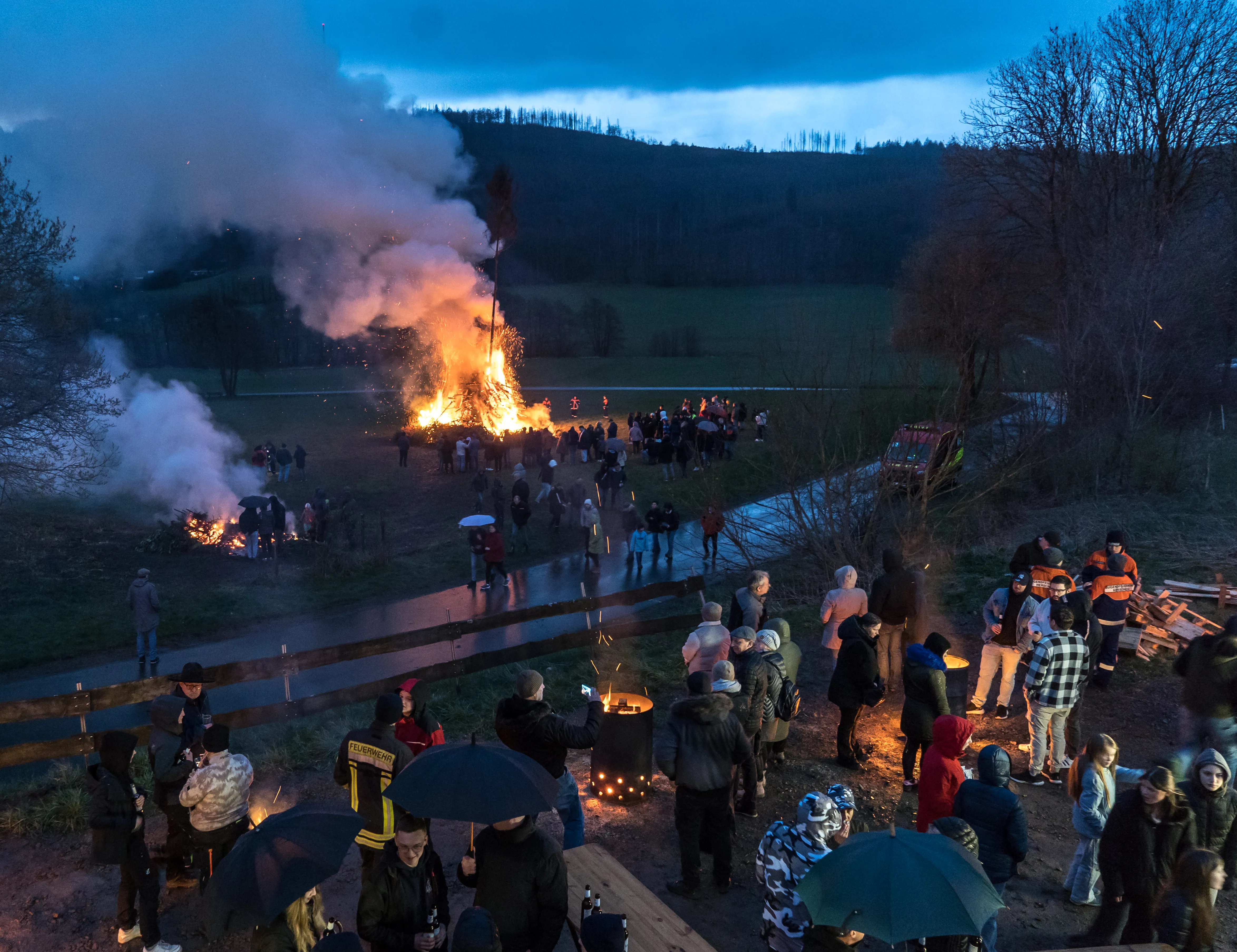 Große Menschenmenge um ein großes Osterfeuer im Freien, im Hintergrund bewaldete Hügel unter blauem Himmel.