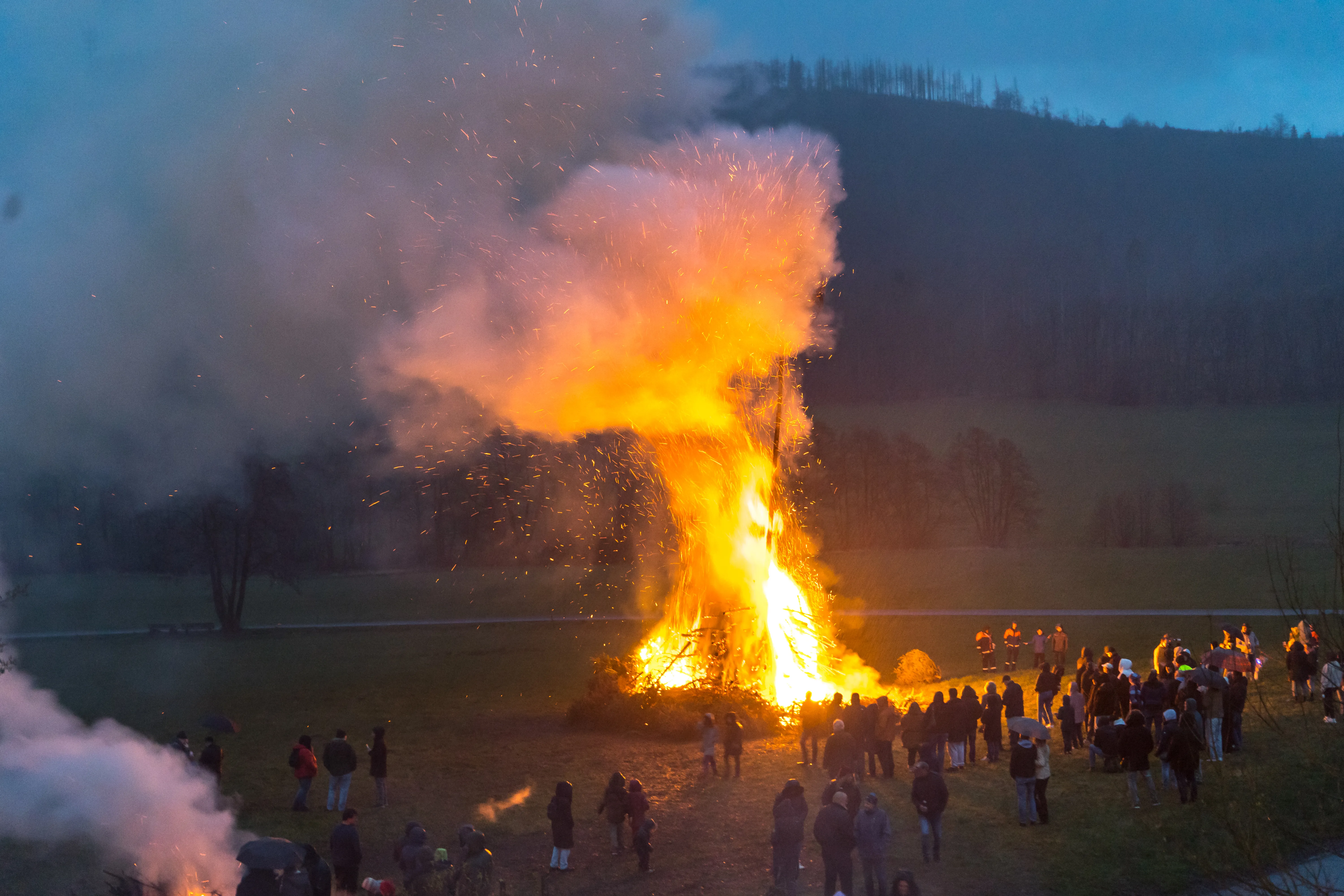 Großes Osterfeuer mit vielen Menschen davor, Rauch und Funken steigen auf. Im Hintergrund eine Hügelkette mit Bäumen.