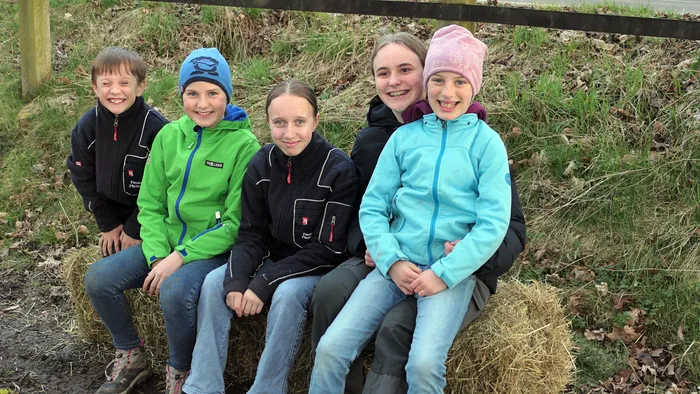Fünf Kinder sitzen lächelnd auf einem Heuballen vor einer grasbewachsenen Böschung.
