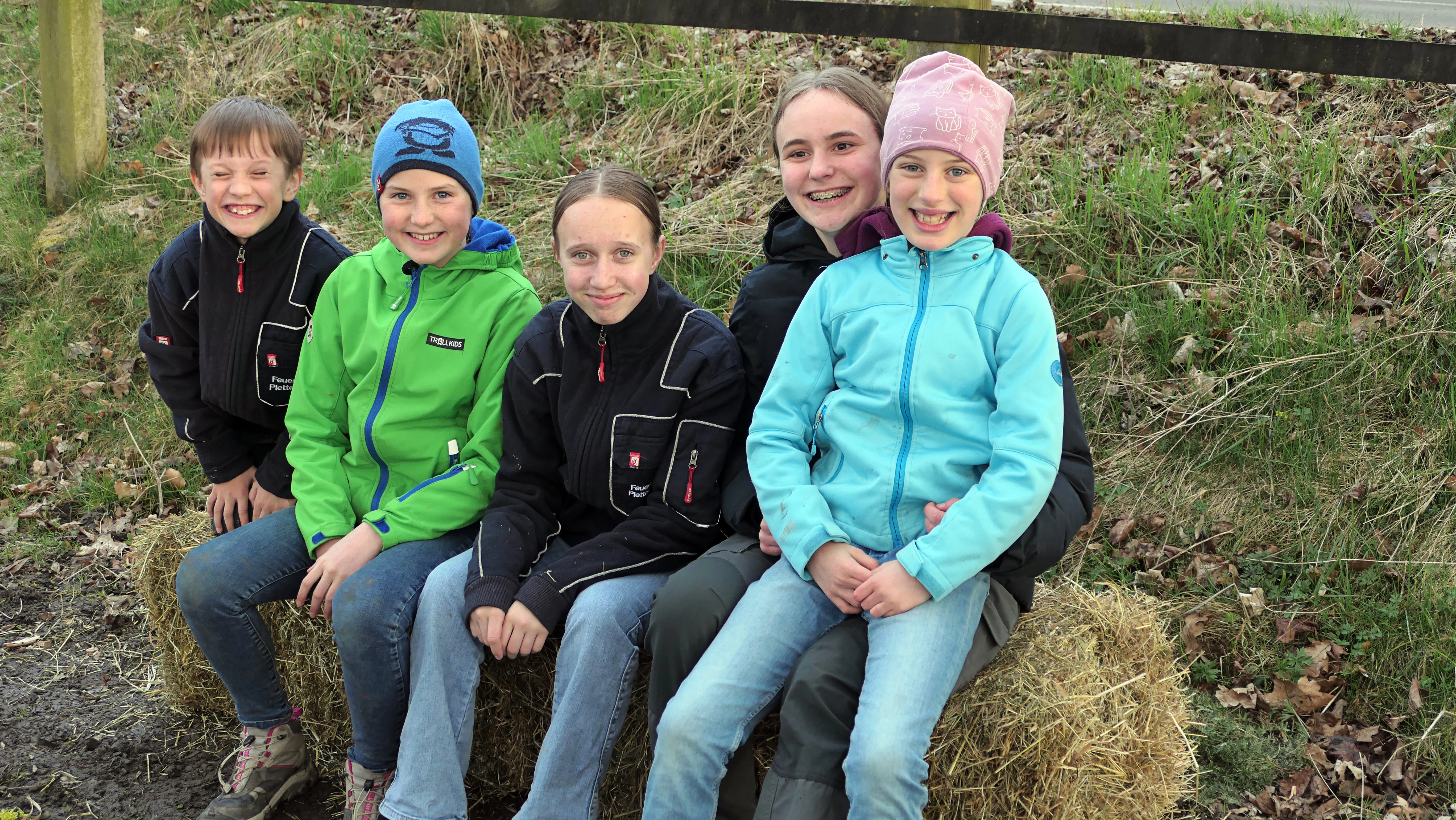 Fünf Kinder sitzen lächelnd auf einem Heuballen vor einer grasbewachsenen Böschung.