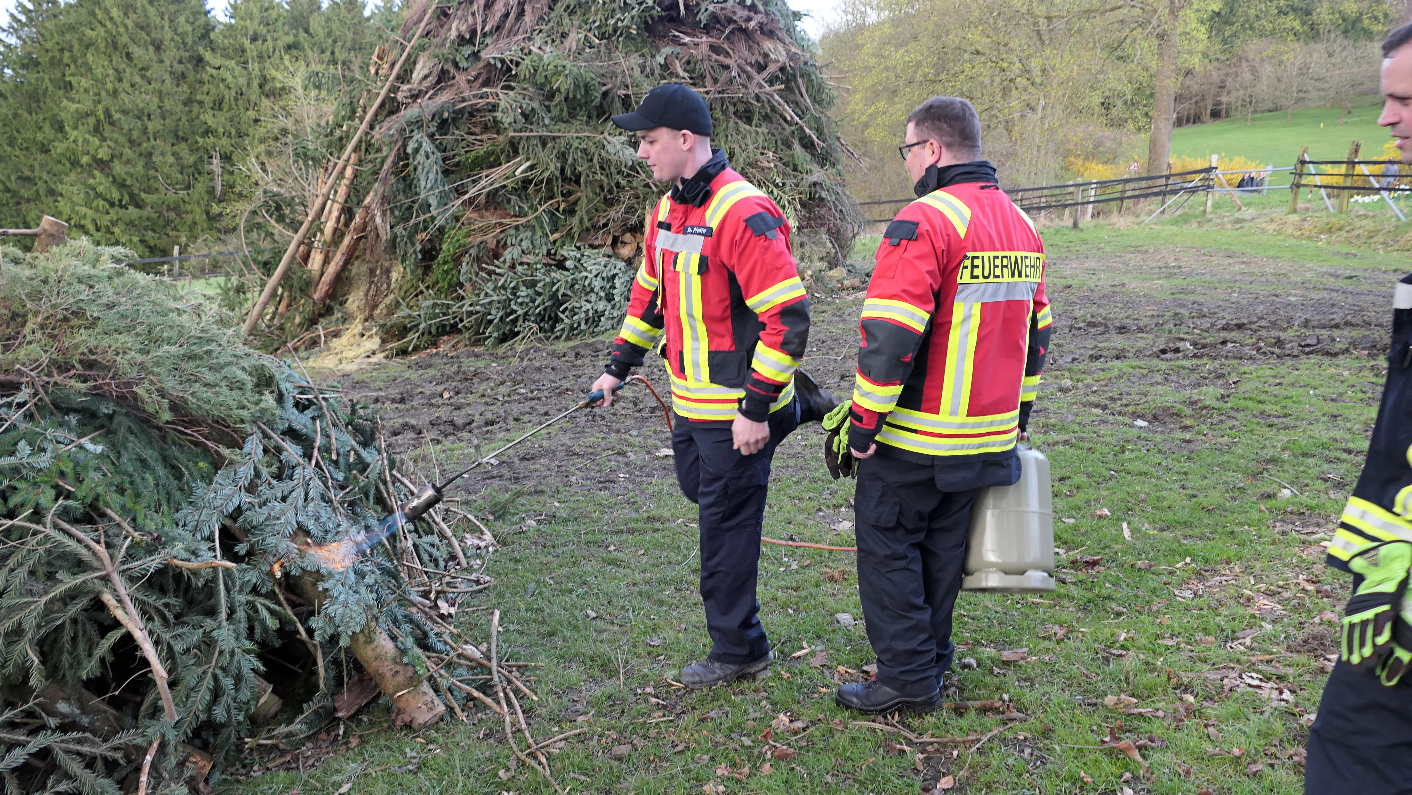 Zwei Feuerwehrleute in voller Montur stehen neben einem großen Haufen aus Ästen und Weihnachtsbäumen, vermutlich zur Überwachung des Osterfeuers.