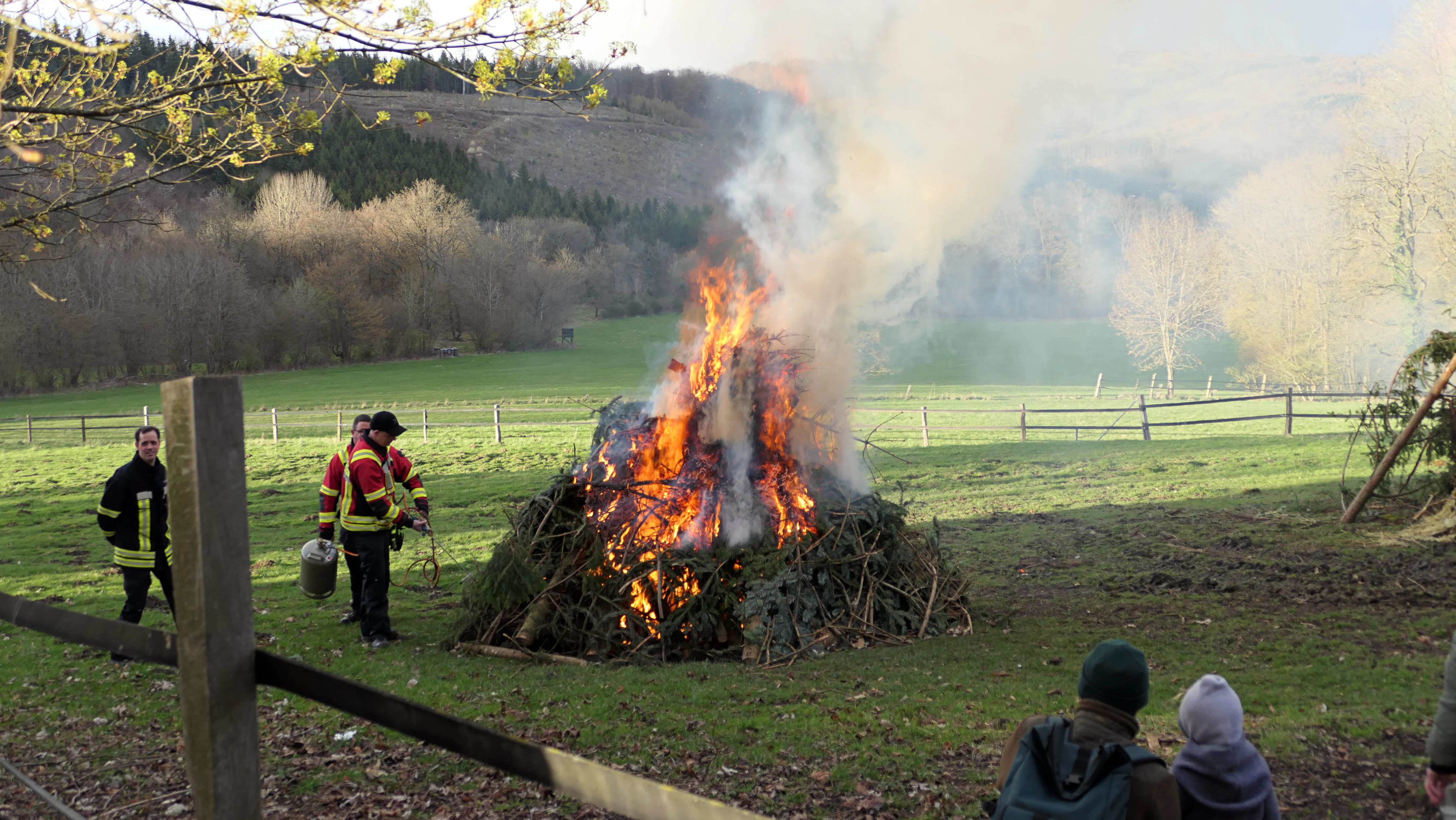 Großes Osterfeuer mit lodernden Flammen und viel Rauch auf einer Wiese, im Hintergrund bewaldete Hügel. Mehrere Personen stehen in der Nähe und beobachten das Feuer.