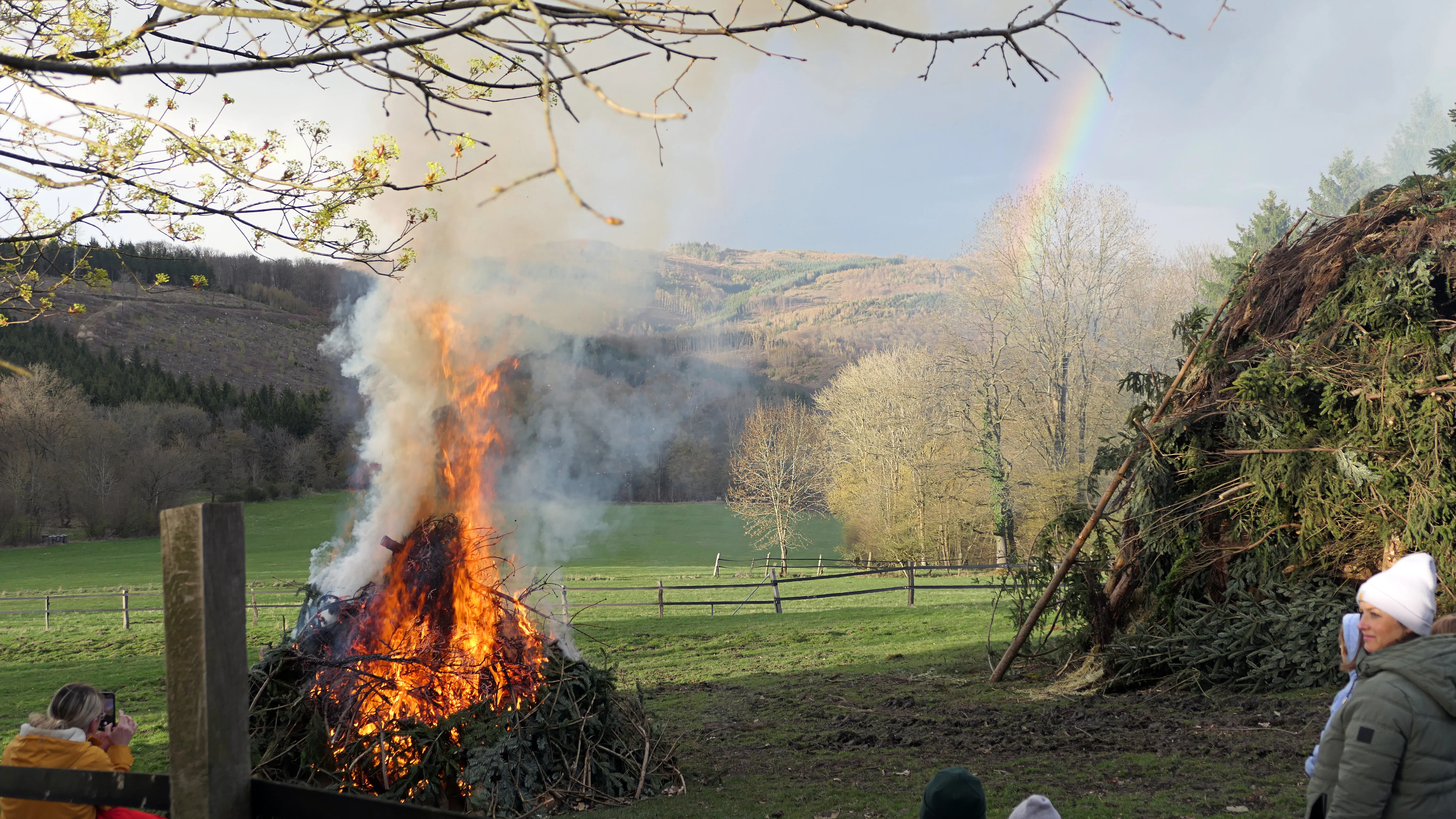 Großes Osterfeuer mit Flammen und Rauch vor hügeliger Landschaft, umgeben von Zuschauern.