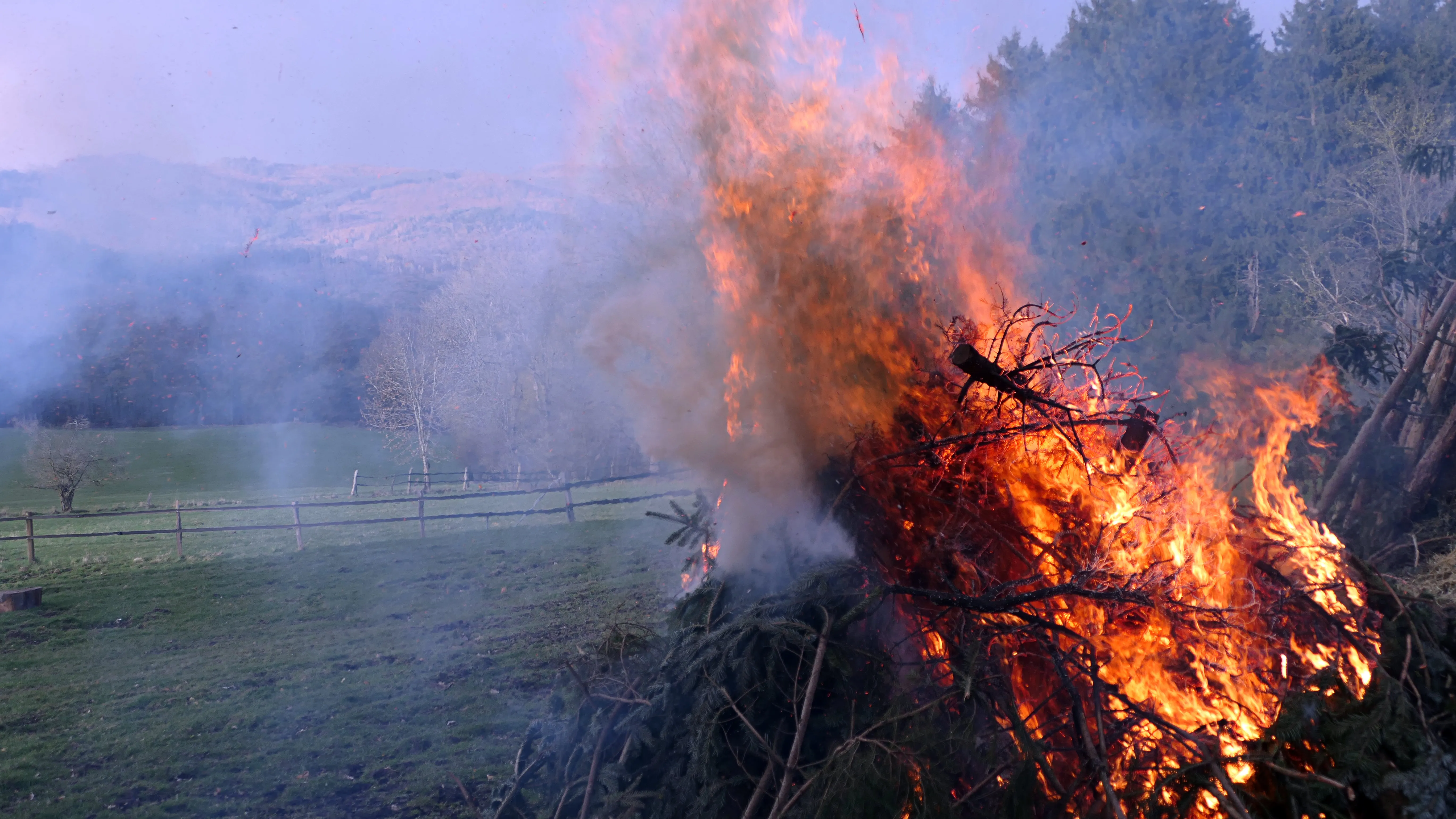 Großes Osterfeuer mit lodernden Flammen und aufsteigendem Rauch auf einer Wiese, im Hintergrund Hügel und Bäume.