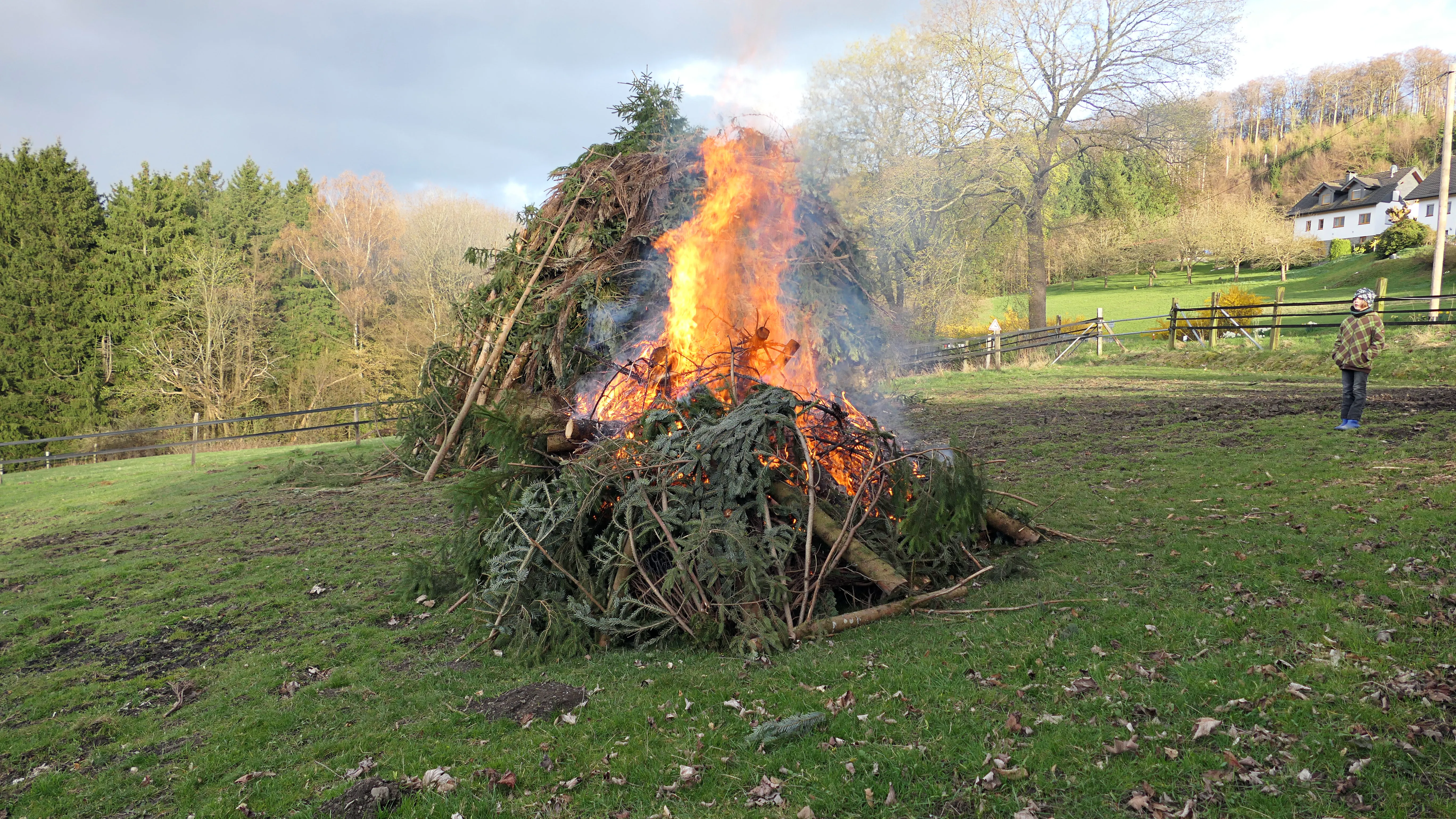 Großes Osterfeuer mit lodernden Flammen und viel Rauch auf einer Wiese, im Hintergrund ein Haus und eine Person.