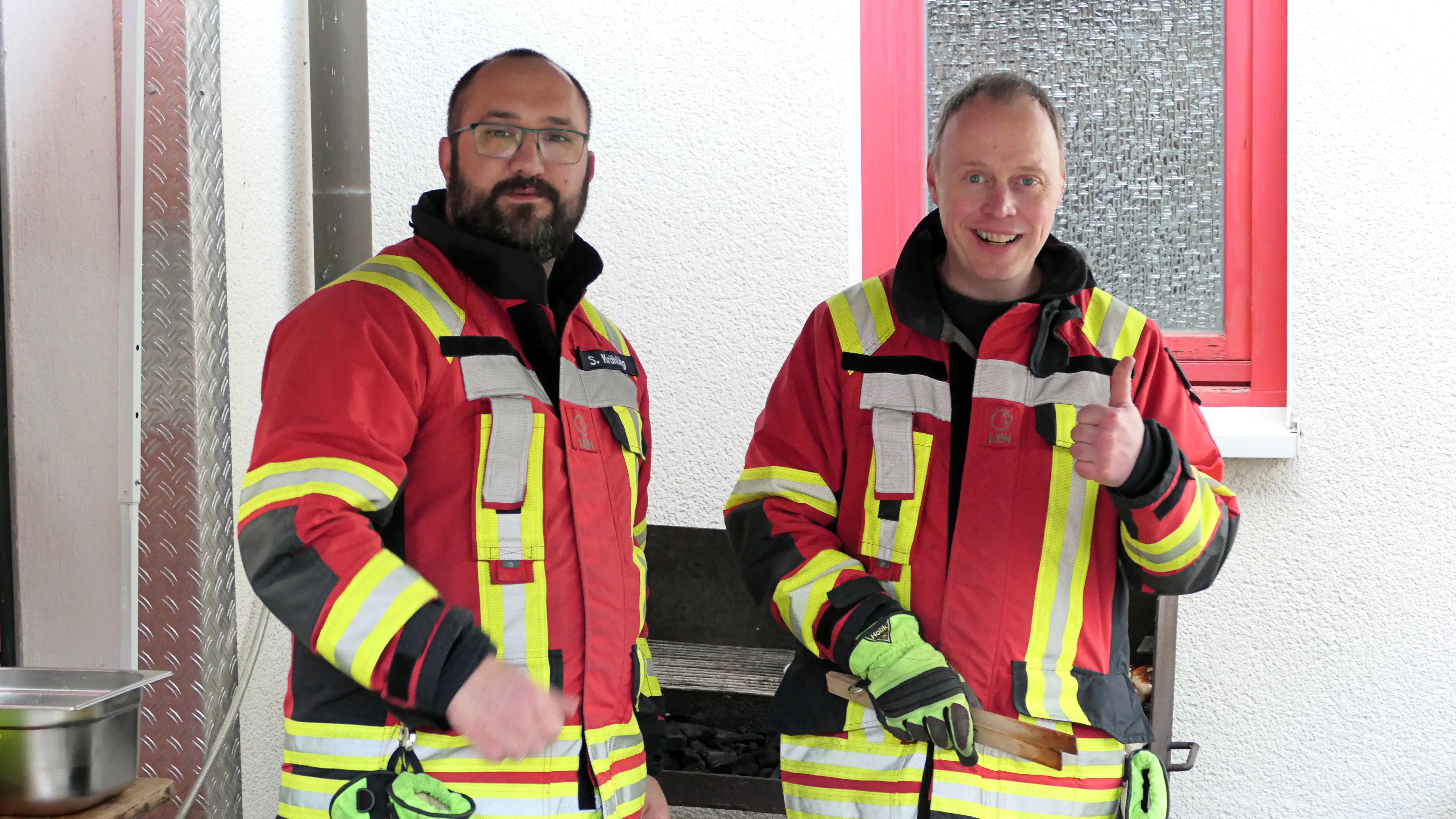 Zwei Feuerwehrleute in voller Montur lächeln in die Kamera, der eine zeigt den Daumen nach oben.