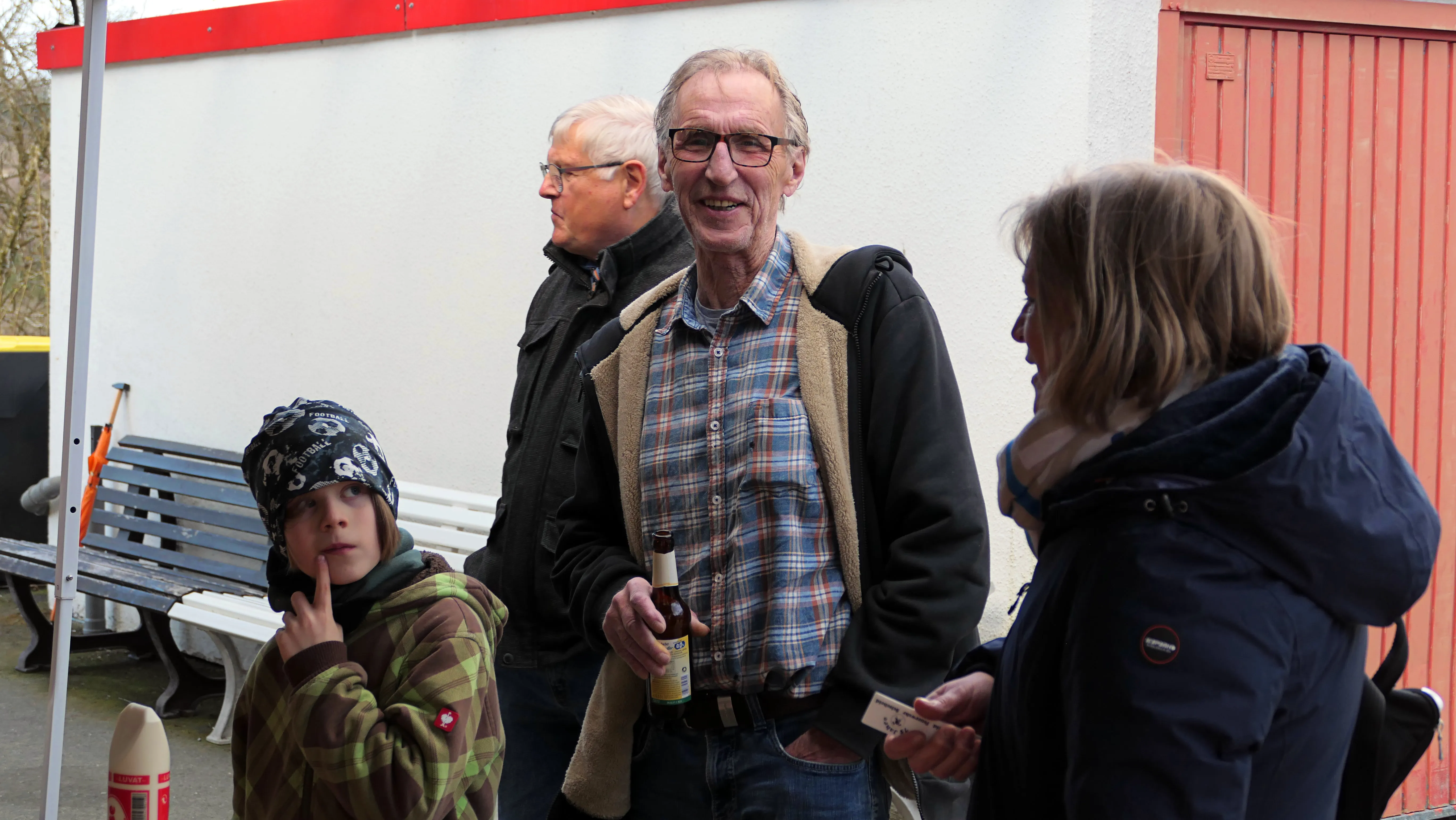 Ein älterer Mann mit Brille lächelt und spricht mit einer Frau, während ein Kind mit Mütze nachdenklich zu ihm aufblickt. Im Hintergrund stehen weitere Personen vor einem roten Gebäude.