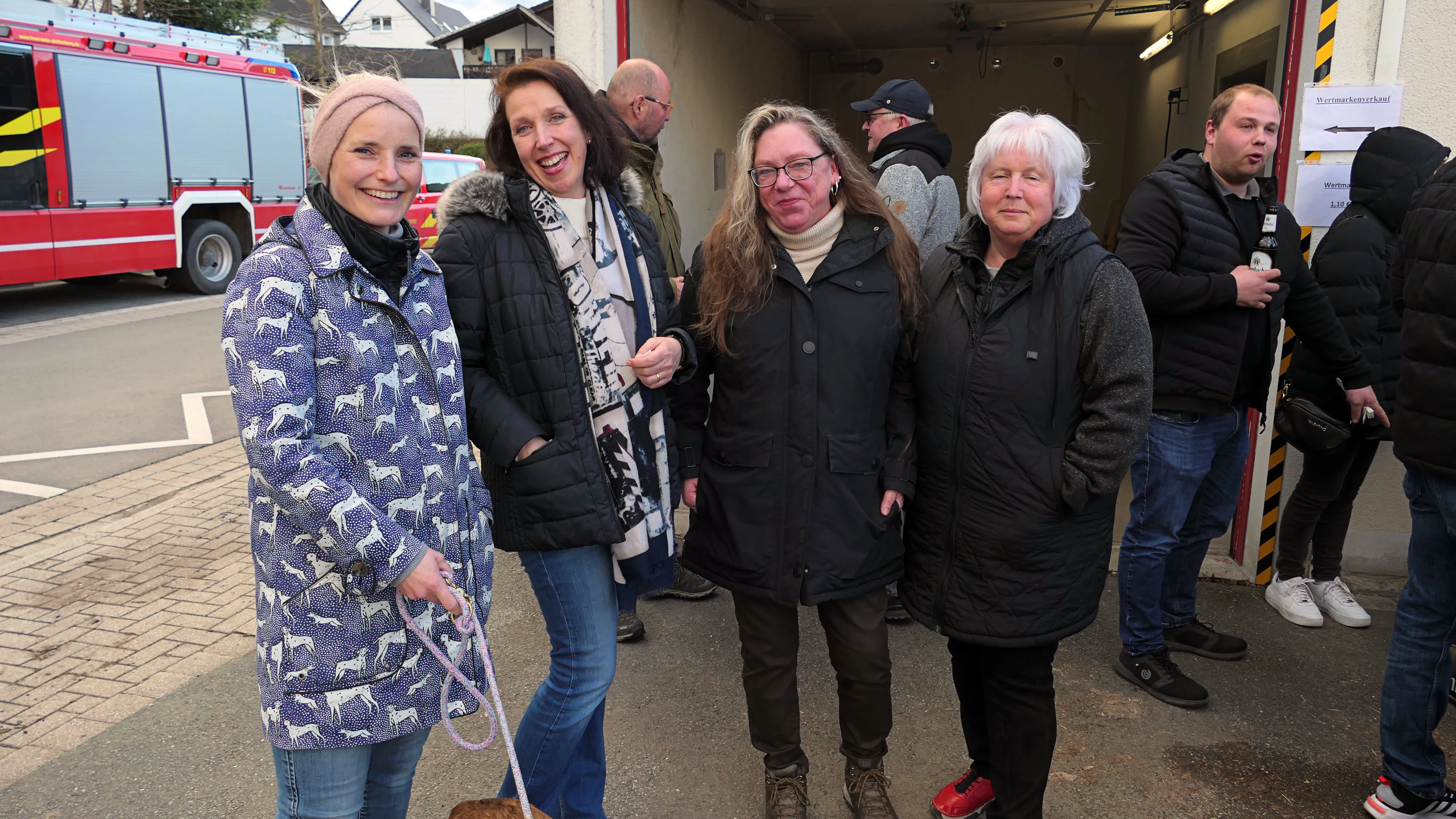 Eine Gruppe von Menschen steht vor einem Feuerwehrhaus, während im Hintergrund ein rotes Feuerwehrauto zu sehen ist.