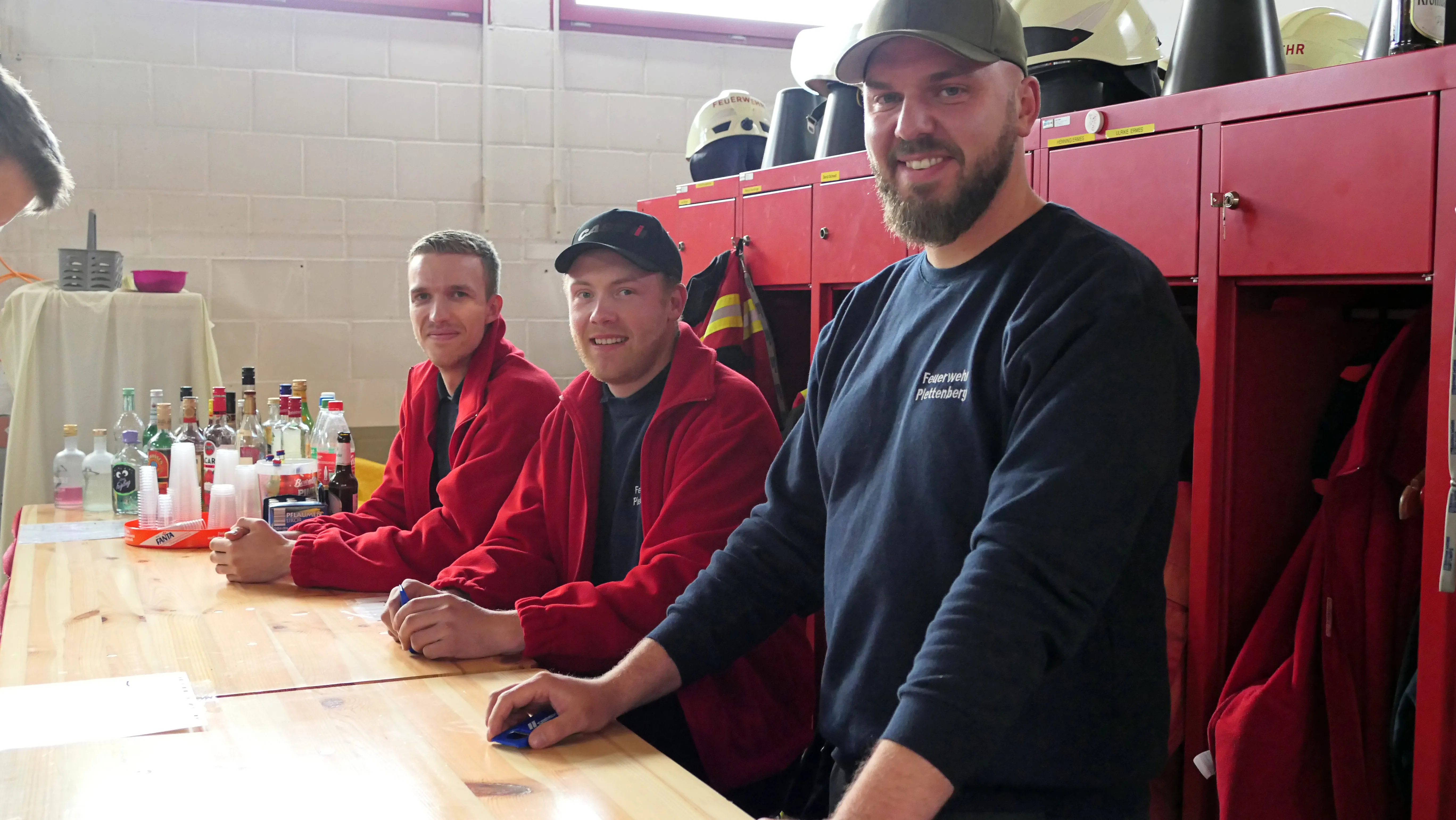 Drei lächelnde Feuerwehrleute in Uniformen stehen an einem Tisch mit Getränken in einem Raum mit roten Schränken und Helmen im Hintergrund.