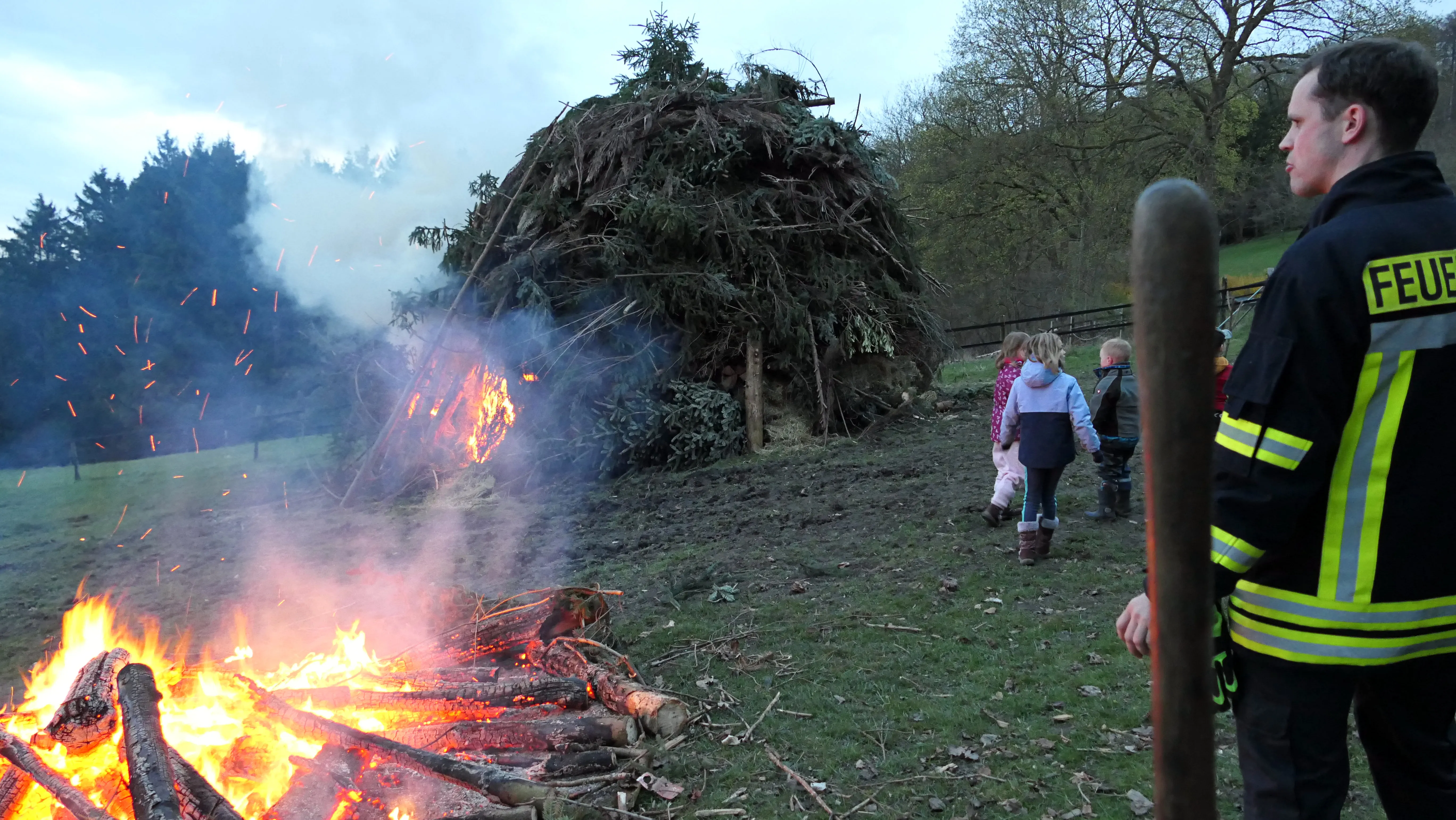 Brennendes Osterfeuer mit großen Holzstapel im Hintergrund, beobachtet von Kindern und einem Feuerwehrmann.