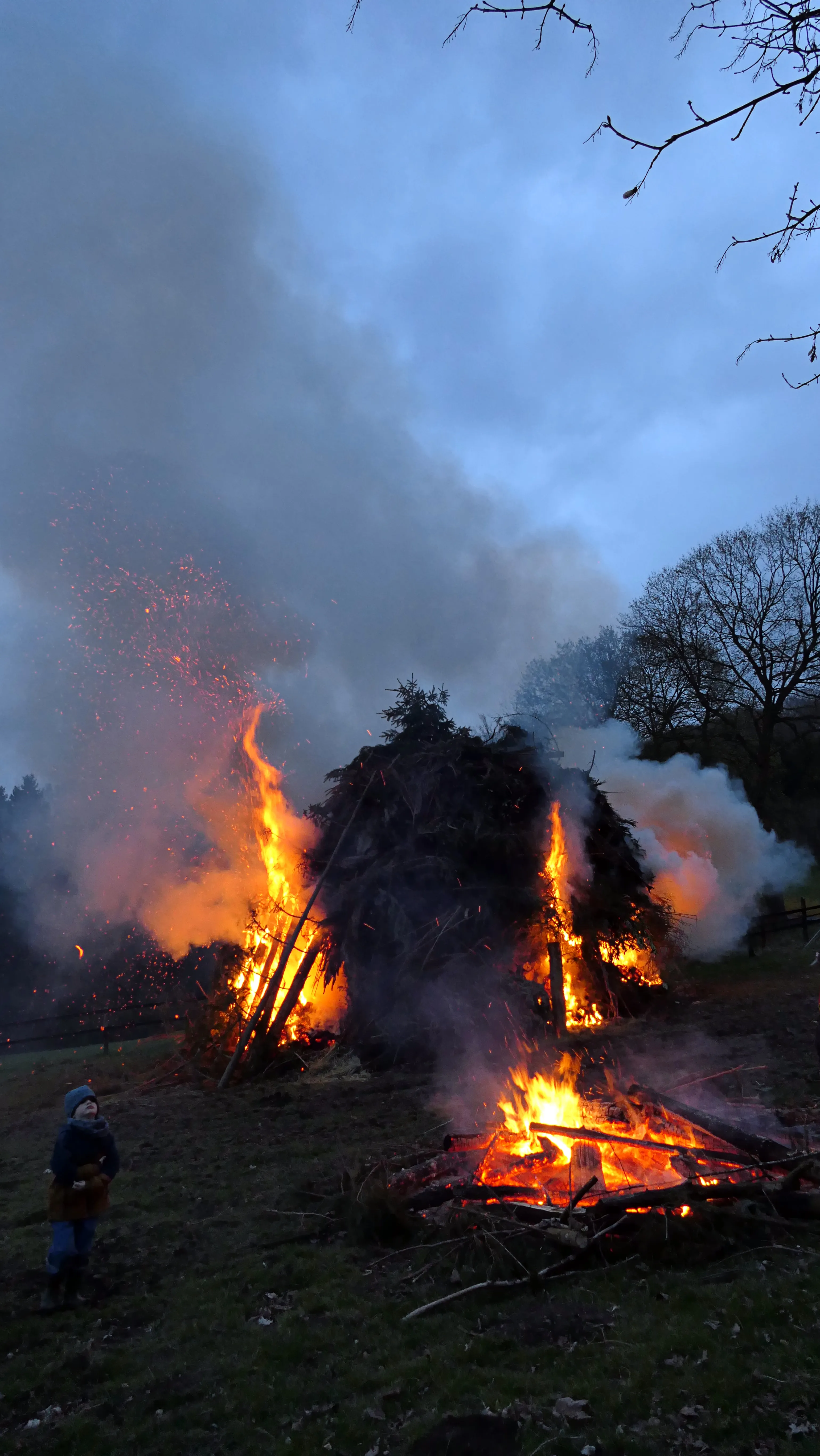 Großes Osterfeuer mit lodernden Flammen und Funkenflug vor bewölktem Himmel, ein Kind steht am Rand.