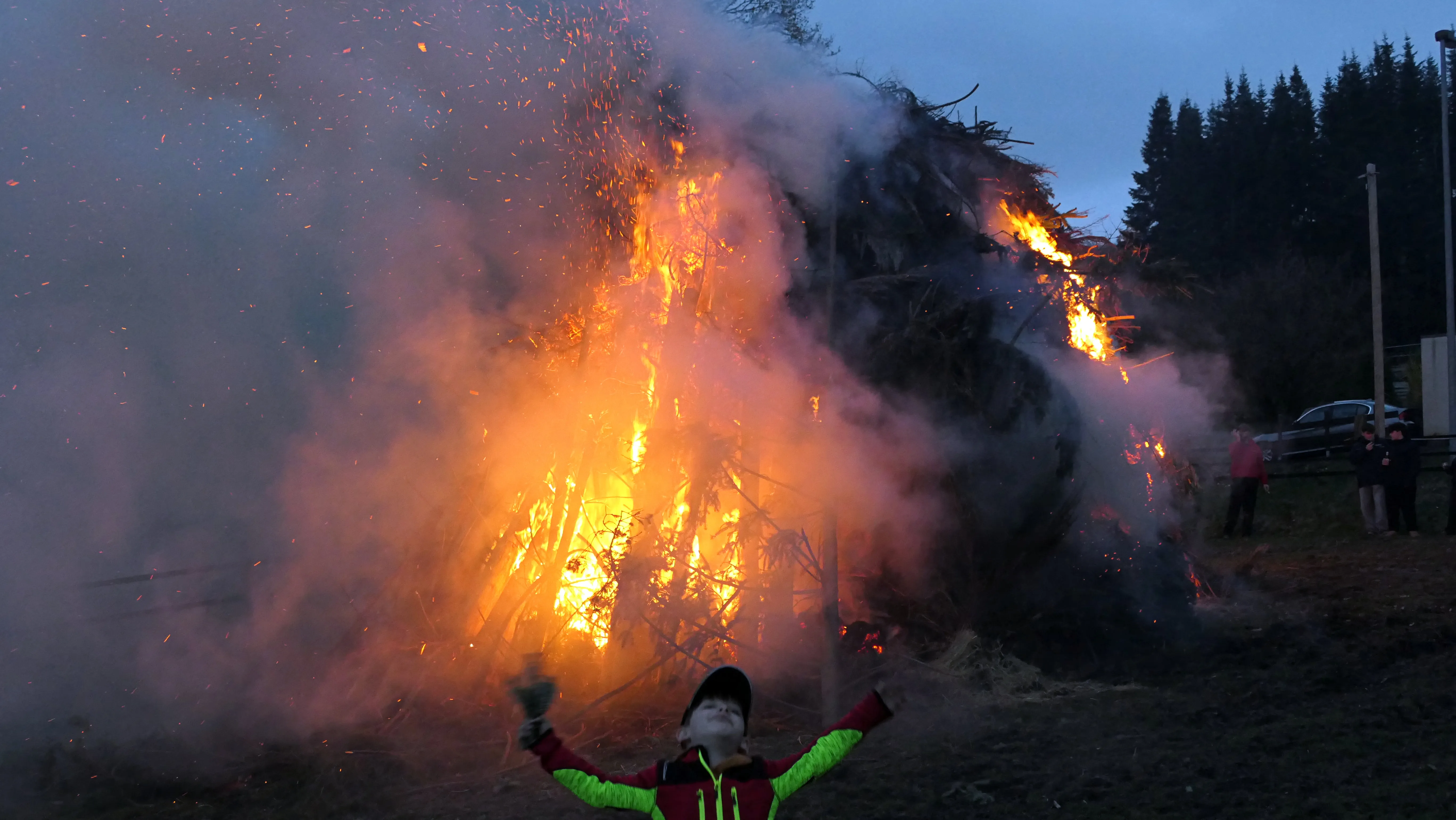 Großes Osterfeuer mit Funkenflug vor dem Hintergrund einer Baumgruppe, im Vordergrund ein Kind mit erhobenen Armen.