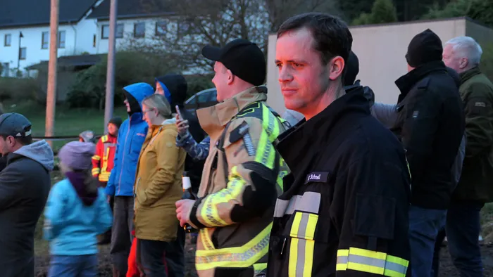 Feuerwehrmann in schwarzer Jacke blickt während eines Osterfeuers in Selscheid in die Ferne, umgeben von weiteren Einsatzkräften und Besuchern.