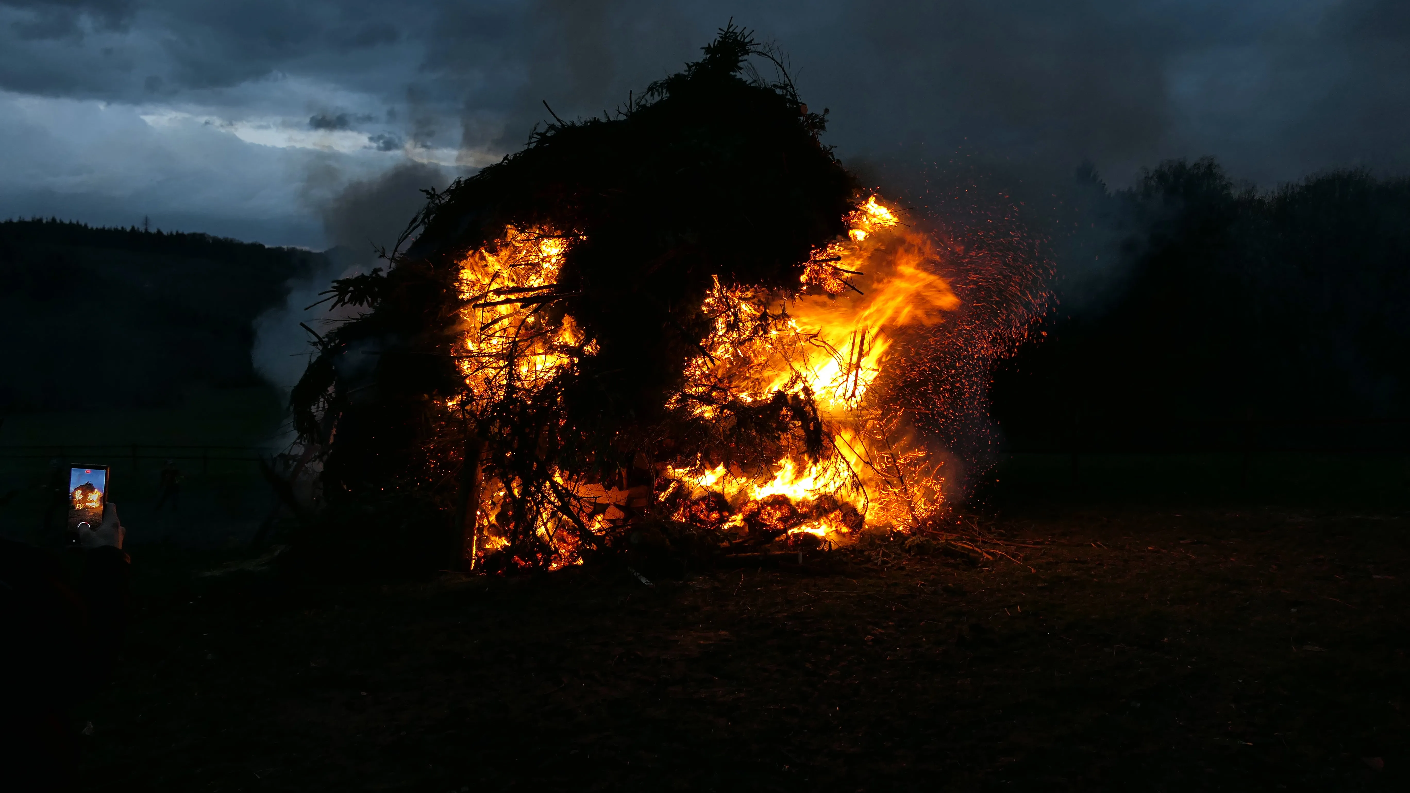 Großes Osterfeuer mit lodernden Flammen und Funkenflug in der Dunkelheit, im Hintergrund eine Person mit Smartphone.