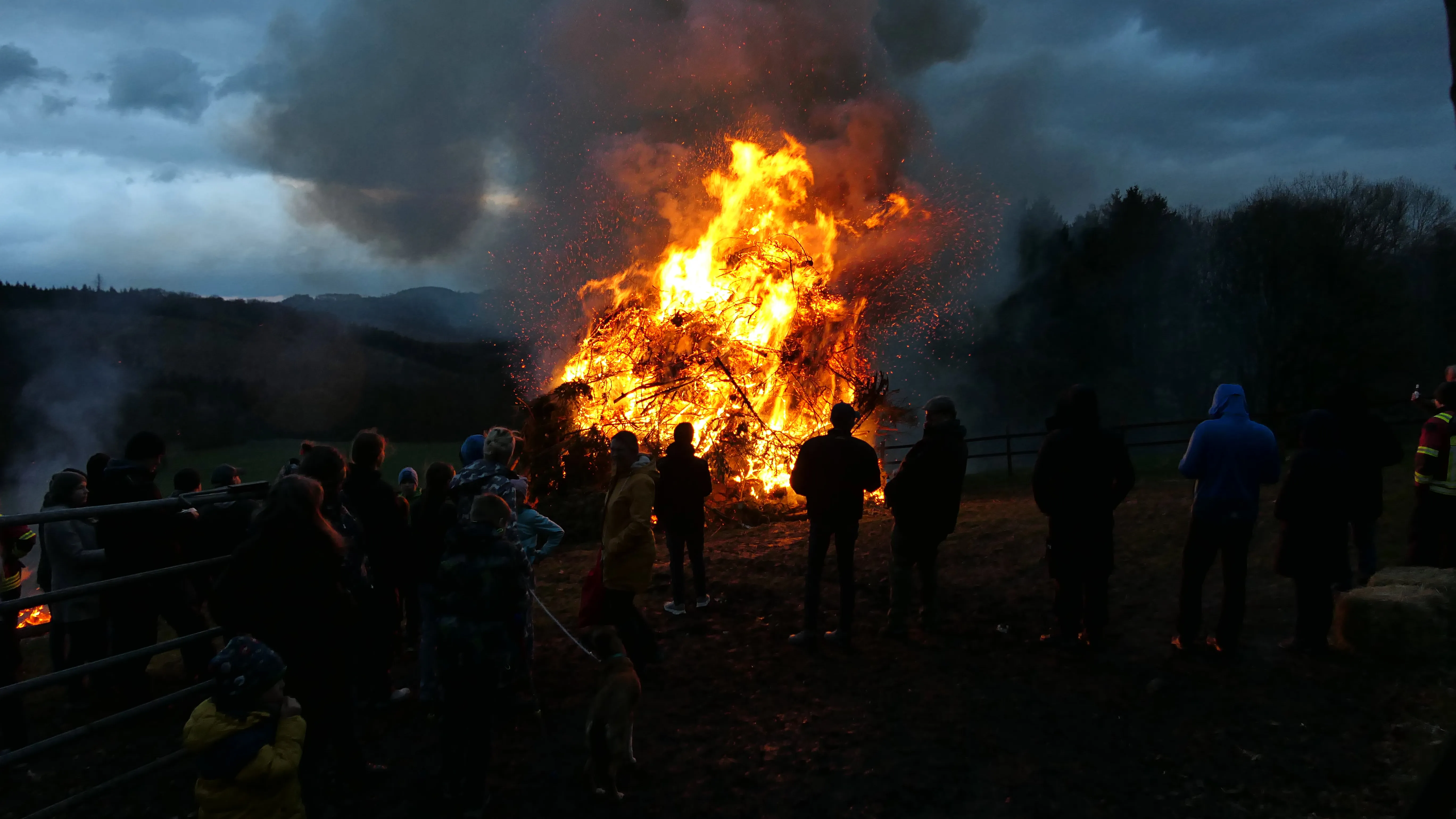 Großes Osterfeuer mit vielen Zuschauern im Abendlicht. Im Hintergrund sind silhouettenhafte Hügel und Bäume zu sehen.