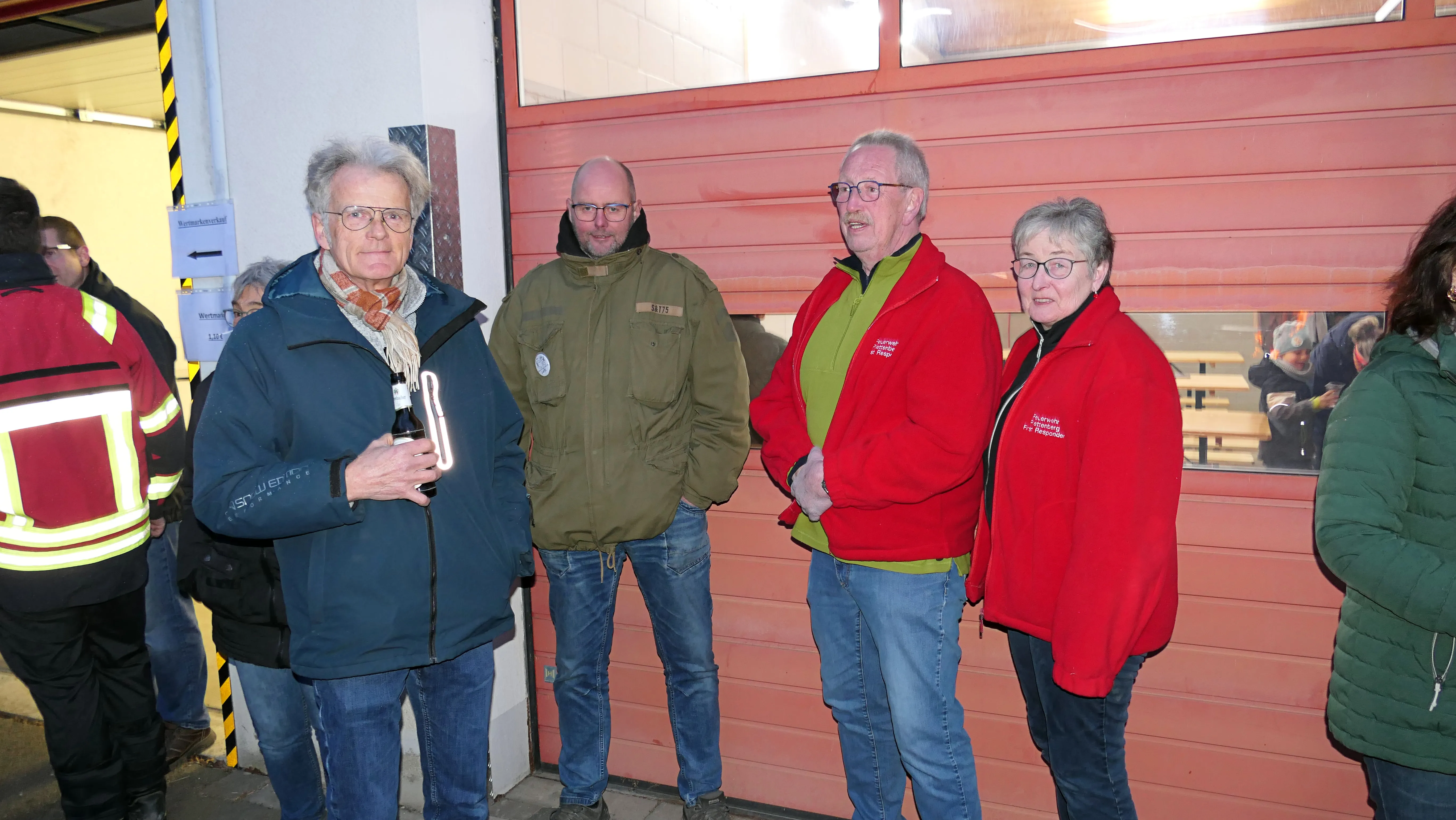 Vier ältere Menschen stehen vor einer geschlossenen Rolltür, während im Hintergrund weitere Personen zu sehen sind. Zwei tragen rote Jacken, die anderen dunklere Winterkleidung.