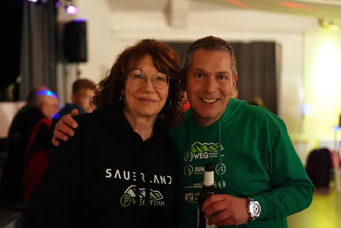 Zwei lächelnde Menschen, eine Frau mit lockigem Haar und ein Mann mit Kurzhaarfrisur, posieren eng nebeneinander. Der Mann trägt ein grünes T-Shirt mit dem Aufdruck „Wander laufen“ und die Frau ein schwarzes Shirt mit dem Logo des P-Wegs.