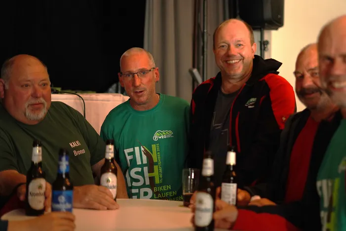 Fünf Männer sitzen an einem Tisch mit Getränken, drei tragen T-Shirts mit Aufdrucken zum P-Weg, einem Wander- und Mountainbike-Event im September 2025.