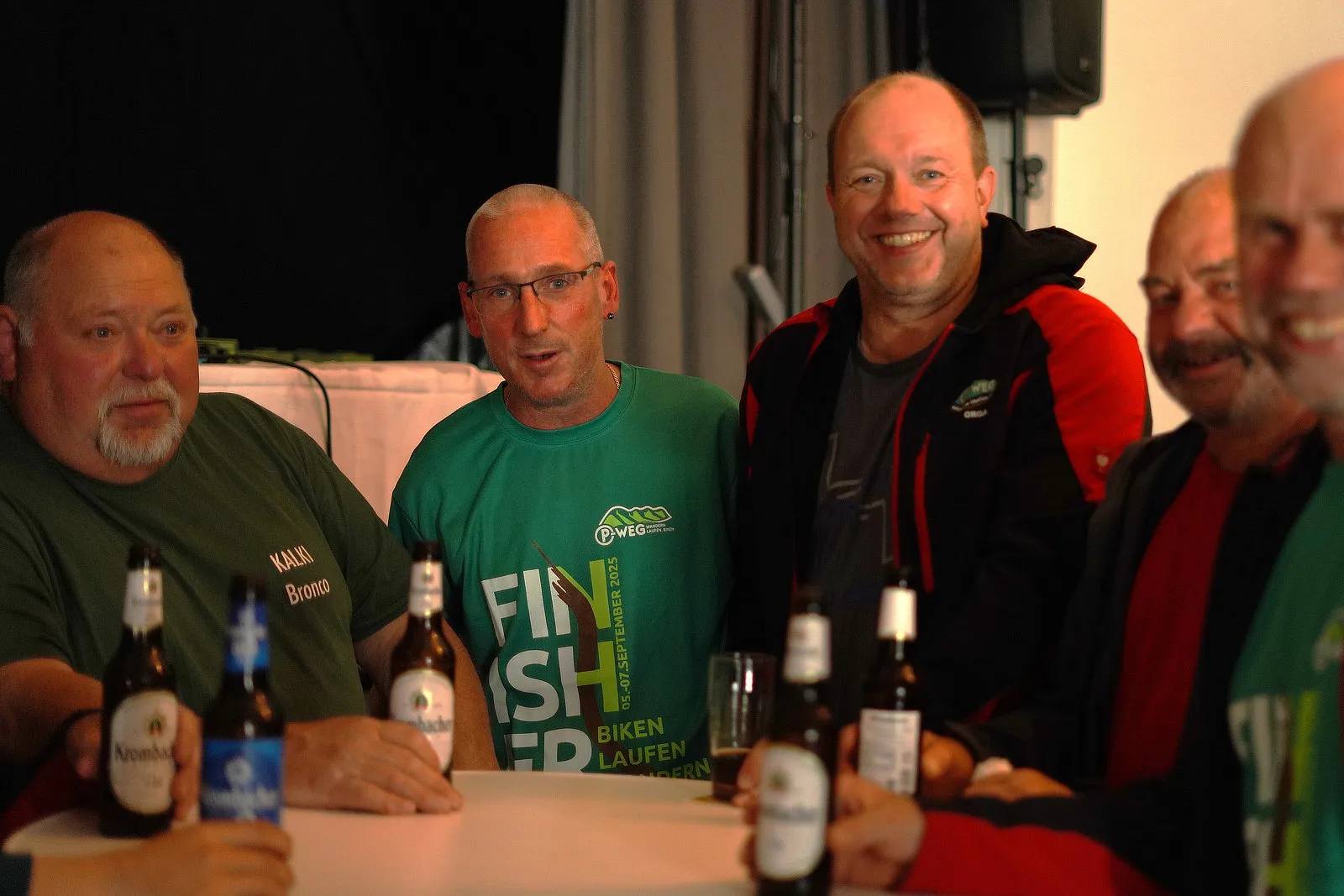 Fünf Männer sitzen an einem Tisch mit Getränken, drei tragen T-Shirts mit Aufdrucken zum P-Weg, einem Wander- und Mountainbike-Event im September 2025.